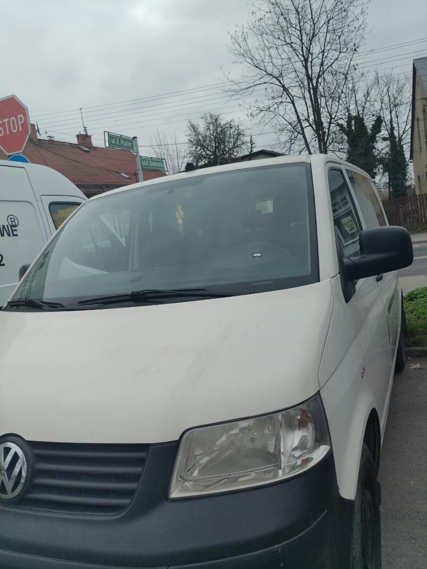 Biały bus Volkswagen Transporter na tle ulicy z tablicami. Widoczny znak STOP i fragment drugiego busa. Pochmurne niebo.