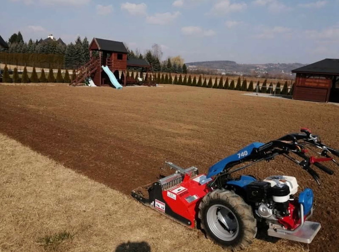 Specjalistyczny glebogryz z czerwonym agregatem spulchnia ziemię na działce z placem zabaw i tujami w tle, przygotowując teren pod przyszły ogród.