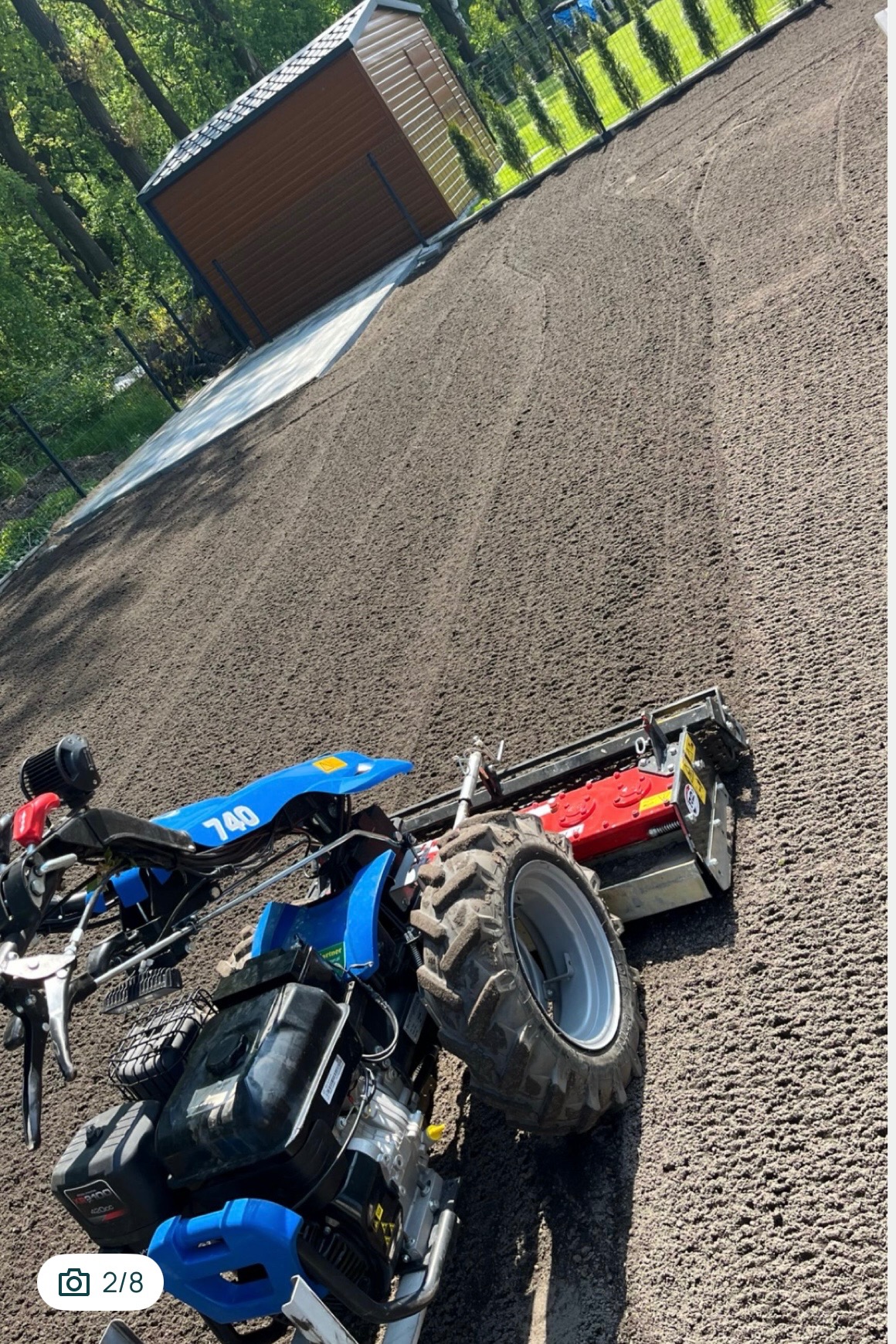 Niwelacja terenu: glebogryzarka frezowa wyrównuje ziemię pod trawnik. W tle domek narzędziowy i zielone tuje wzdłuż ogrodzenia. Widok z góry, lekki kąt.