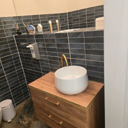 Krzysztofa Płosaj-Sandzewicz - Nowoczesna łazienka: biała umywalka nablatowa z złotą armaturą na drewnianej szafce, ciemne płytki na ścianie, marmurowe płytki podłogowe. Minimalistyczny design.