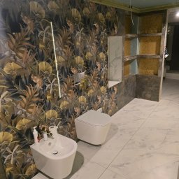 Krzysztofa Płosaj-Sandzewicz - Nowoczesna łazienka w trakcie wykończenia: biała armatura (bidet i toaleta) na tle marmurowej podłogi i ściany z motywem roślinnym. Widoczne półki w zabudowie.