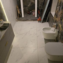 Krzysztofa Płosaj-Sandzewicz - Białe płytki imitujące marmur w łazience w Kamiennej Górze. Widoczna toaleta, bidet i szafka pod umywalkę. W tle narzędzia i materiały budowlane.