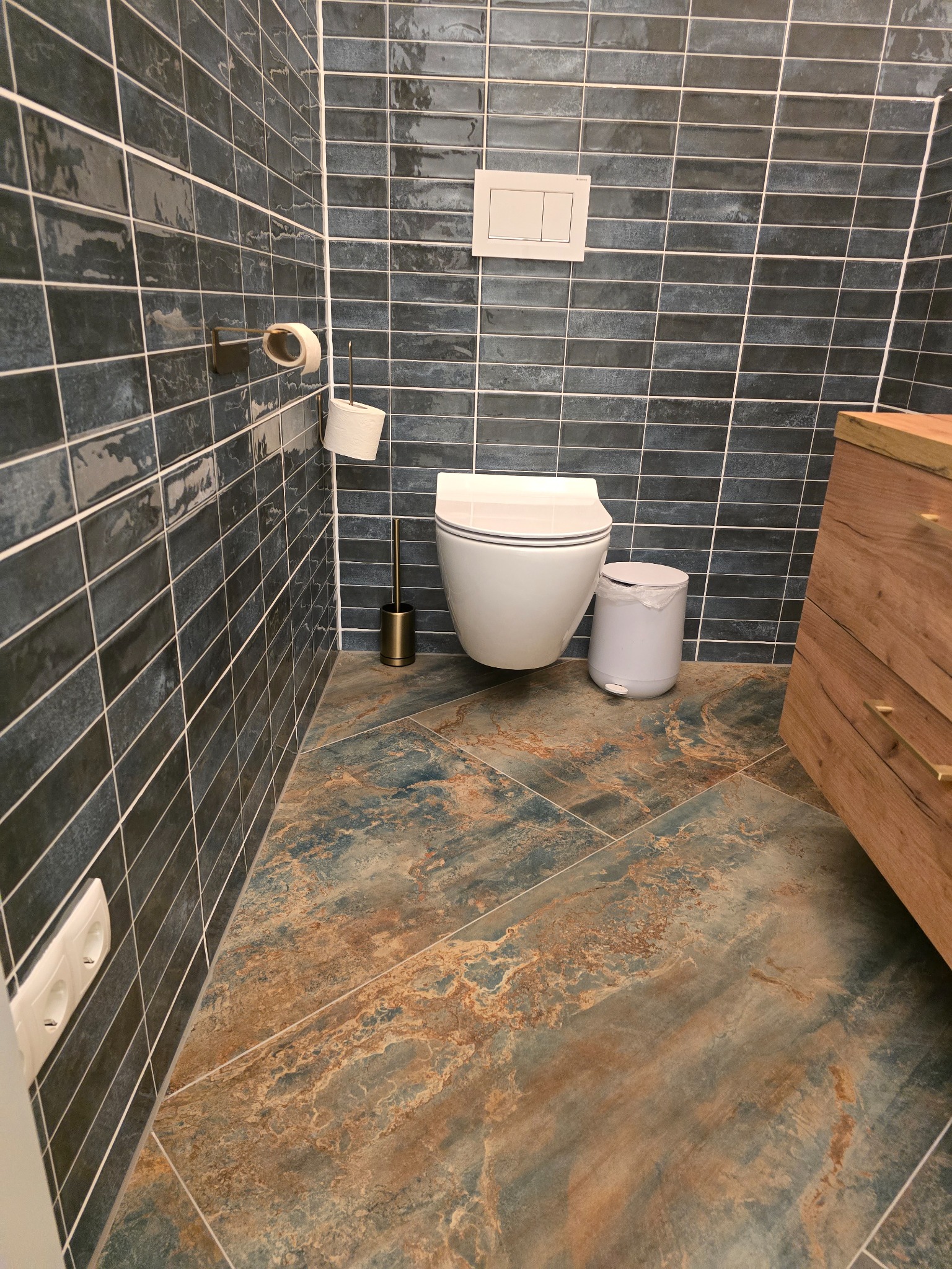 Nowoczesna łazienka z ciemnymi, podłużnymi płytkami na ścianach i wzorzystą, brązowo-niebieską podłogą. Widoczna toaleta, kosz na śmieci i drewniana szafka.