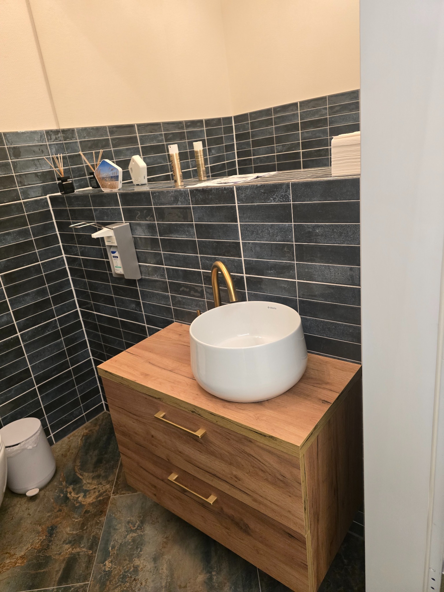 Nowoczesna łazienka: biała umywalka nablatowa z złotą armaturą na drewnianej szafce, ciemne płytki na ścianie, marmurowe płytki podłogowe. Minimalistyczny design.