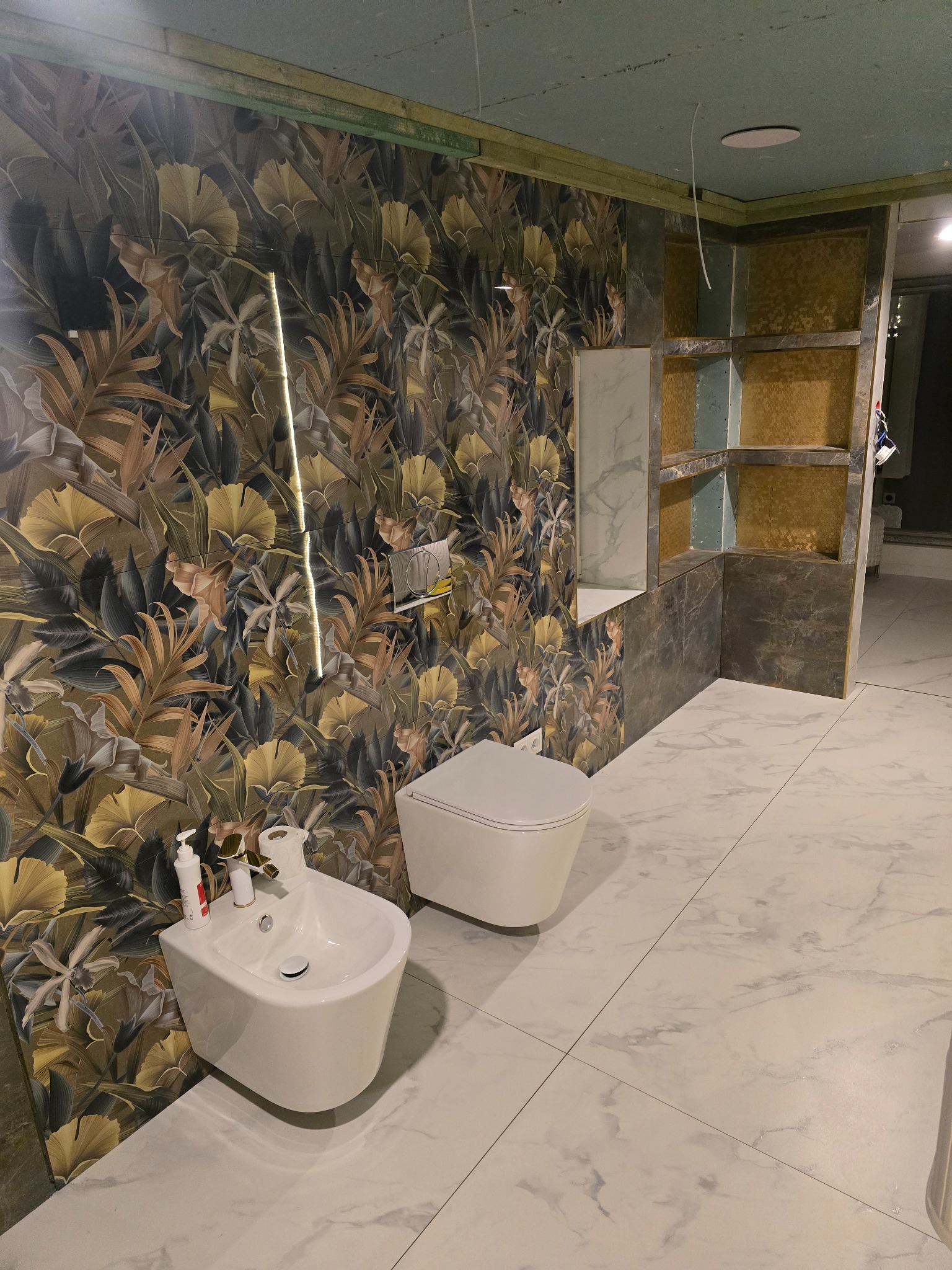 Nowoczesna łazienka w trakcie wykończenia: biała armatura (bidet i toaleta) na tle marmurowej podłogi i ściany z motywem roślinnym. Widoczne półki w zabudowie.