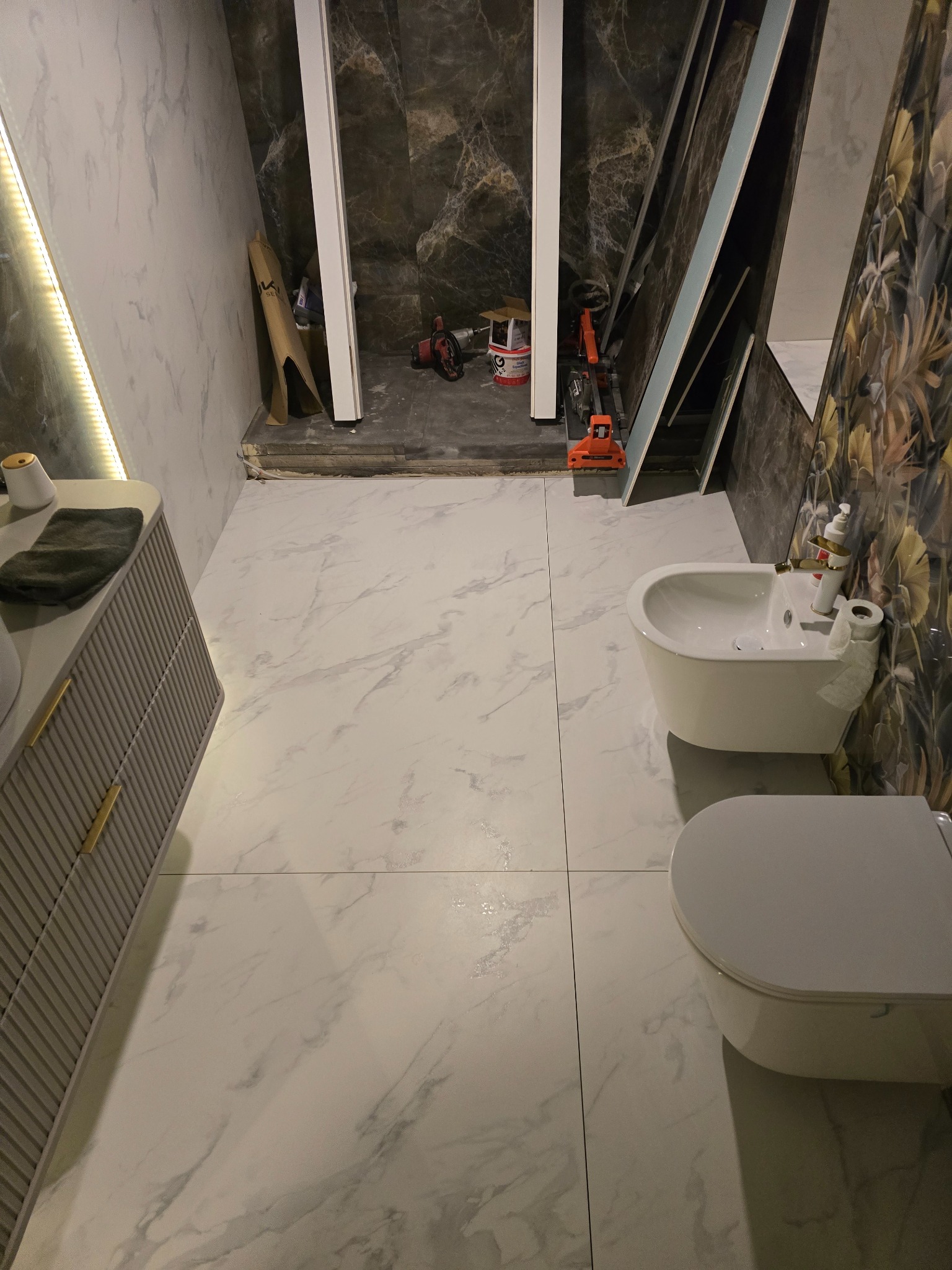 Białe płytki imitujące marmur w łazience w Kamiennej Górze. Widoczna toaleta, bidet i szafka pod umywalkę. W tle narzędzia i materiały budowlane.