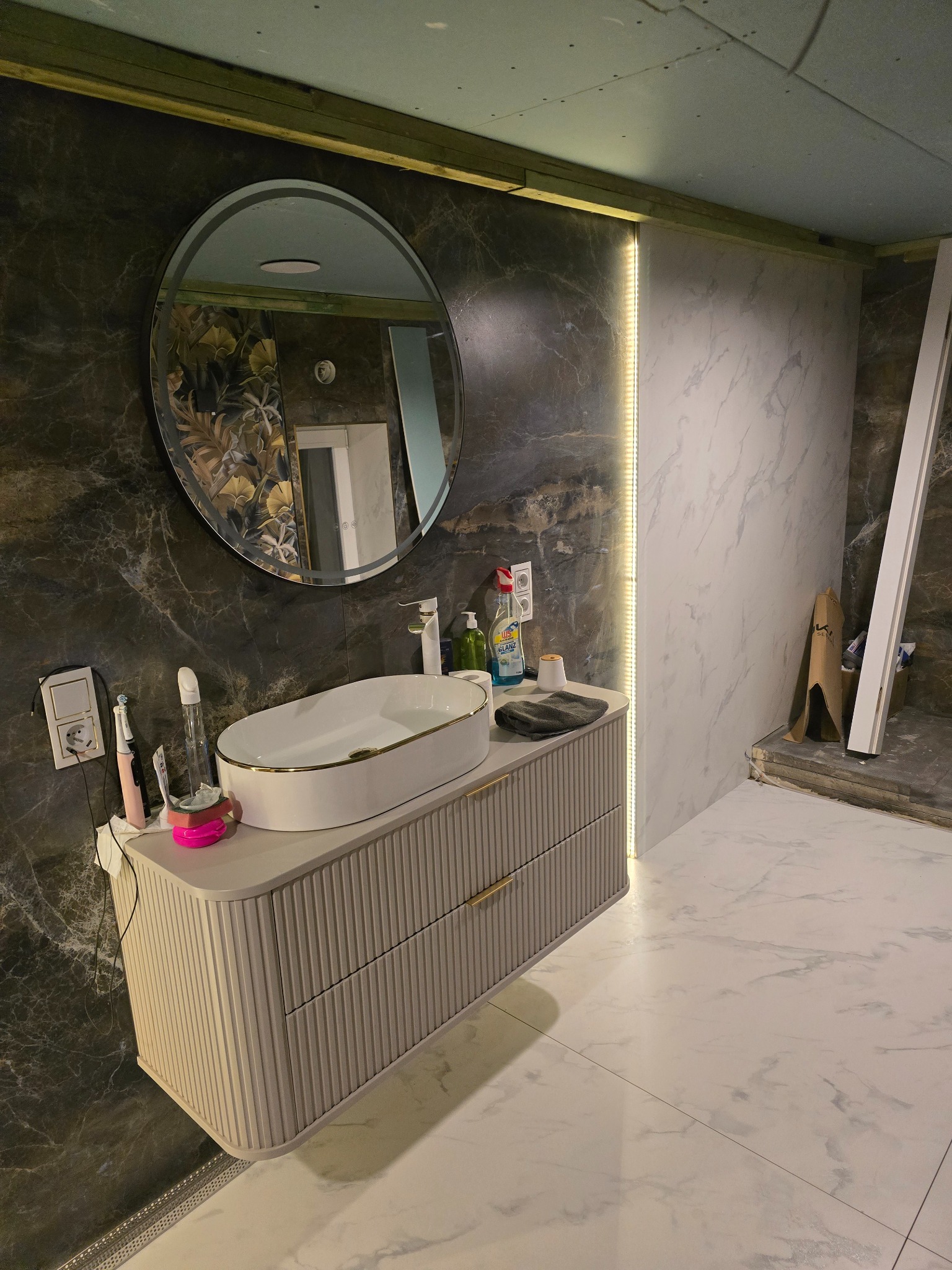 Nowoczesna łazienka w trakcie wykończenia: marmurowe ściany, owalne lustro, biała umywalka z szafką. Widoczne elementy instalacji i płyty karton-gips na suficie.