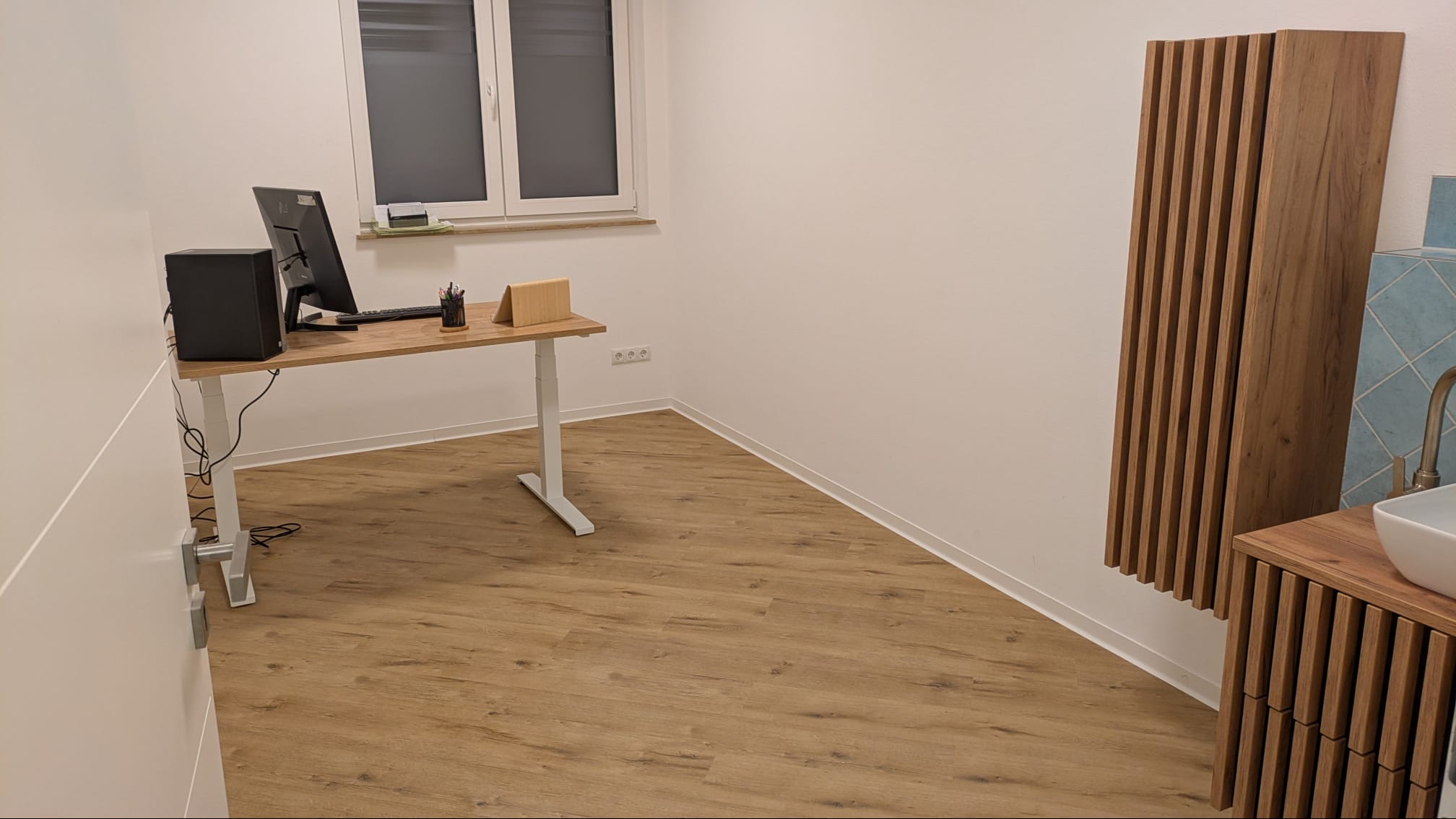 Nowoczesne wnętrze z drewnianą podłogą, biurkiem z komputerem i dekoracyjnym panelem ściennym z drewnianych listew w łazience. Minimalistyczny design.