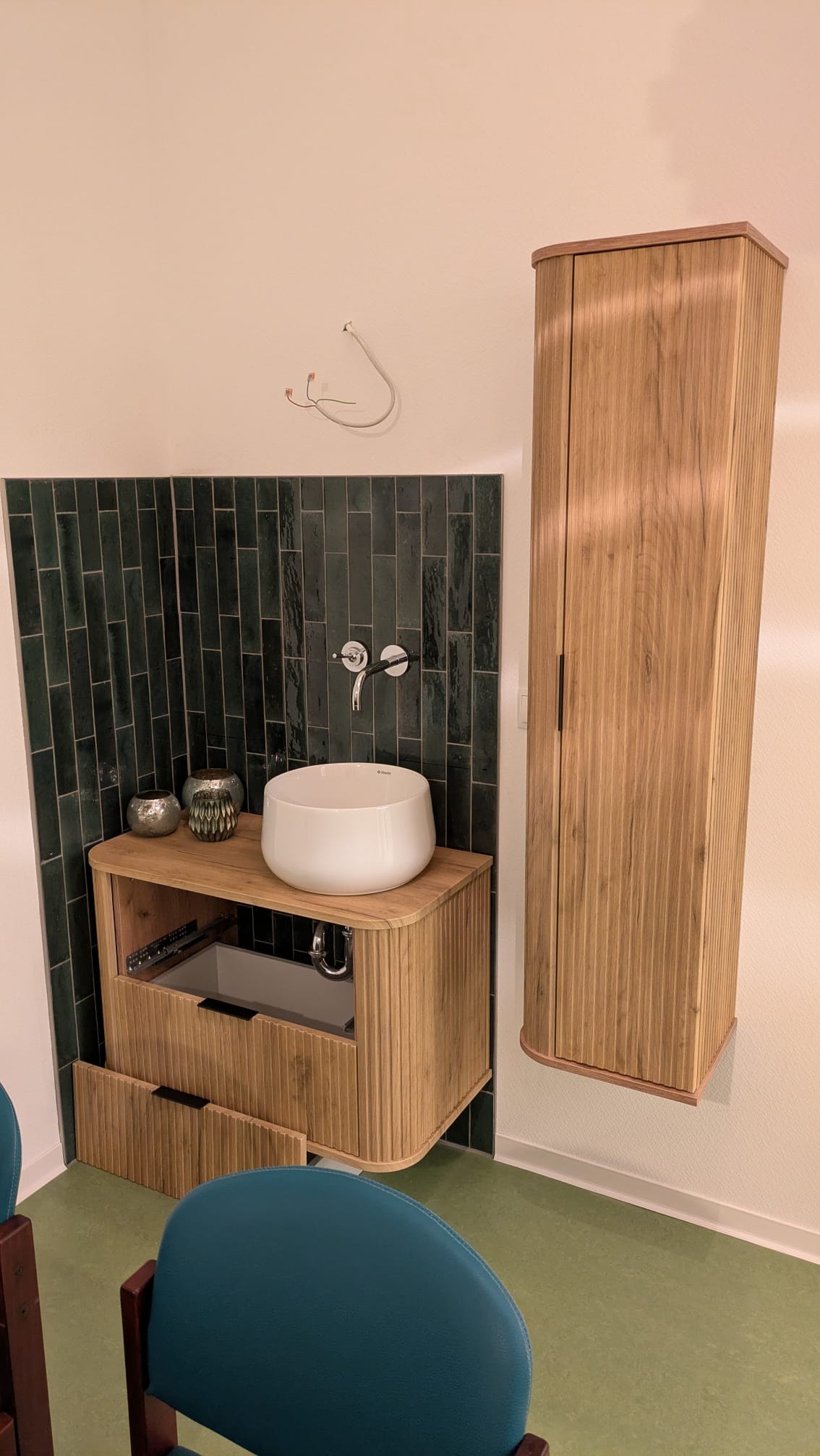 Nowoczesna łazienka z umywalką nablatową, szafką z lamelami i ciemnozielonymi płytkami. Widoczne niedokończone podłączenie elektryczne.