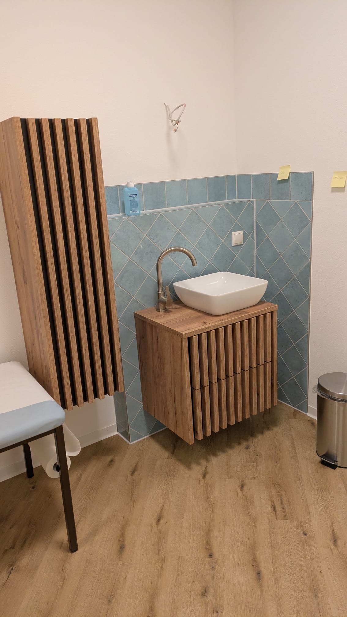 Nowoczesna łazienka po remoncie: drewniana szafka z umywalką nablatową, dekoracyjne panele ścienne i podłoga z paneli w odcieniach drewna. Błękitne płytki w tle.