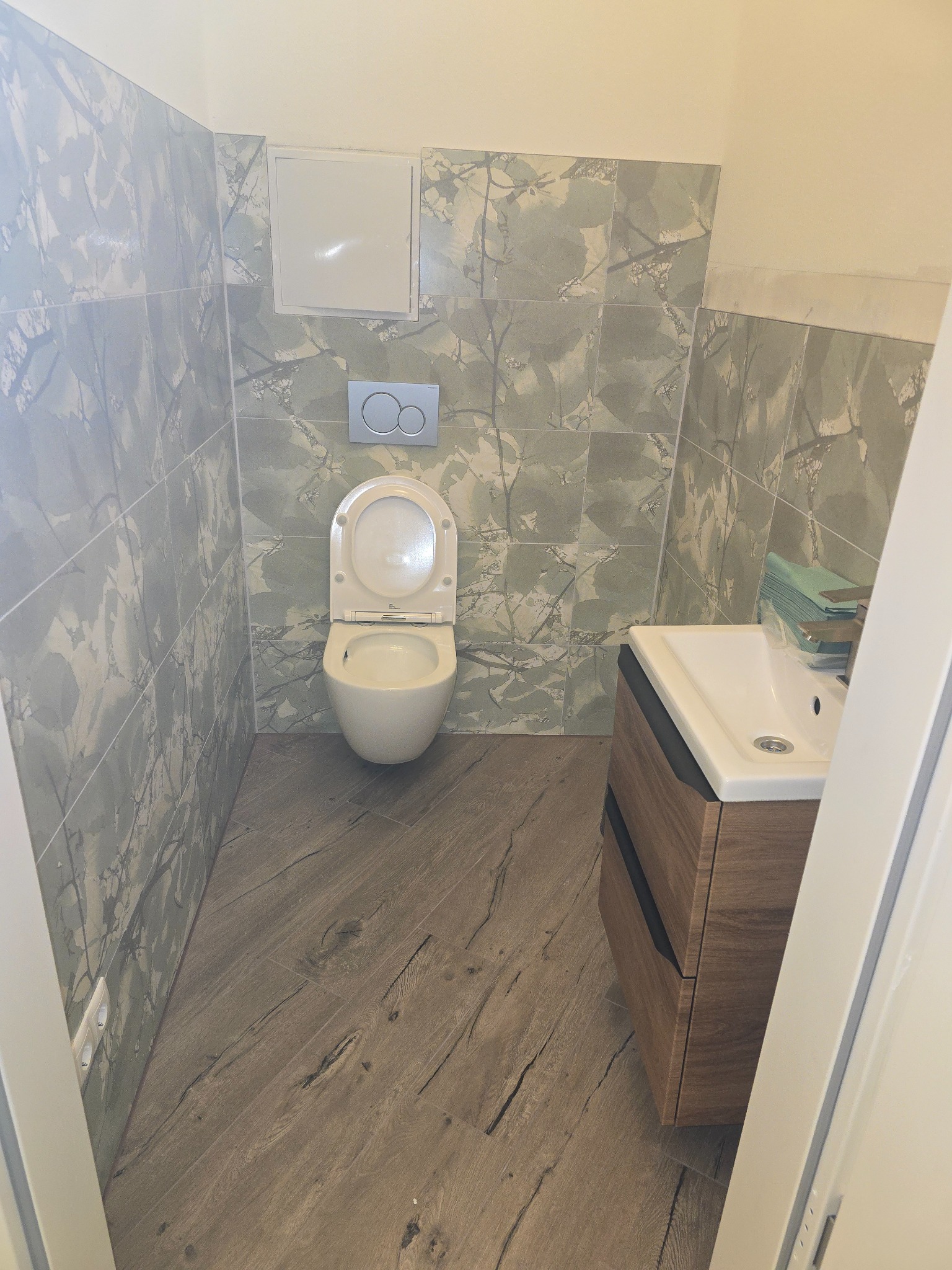 Ukończona łazienka z toaletą, umywalką z szafką w odcieniach brązu i szaro-białymi płytkami z motywem liści na ścianach i podłodze imitującej drewno.