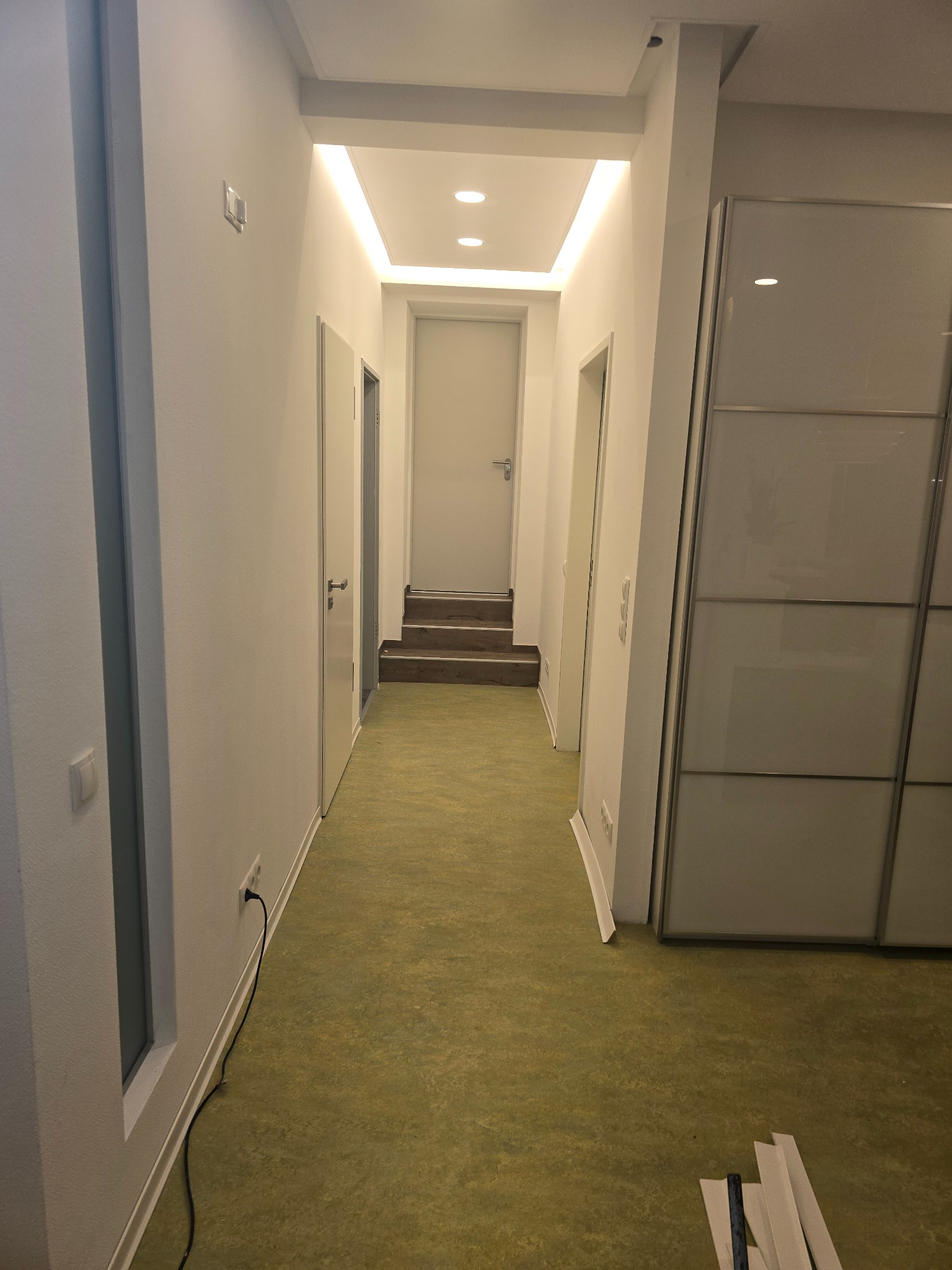 Korytarz po remoncie z oświetleniem LED, zieloną podłogą i białą zabudową szafy. Widoczne listwy przypodłogowe i gniazdko elektryczne. Minimalistyczny design.