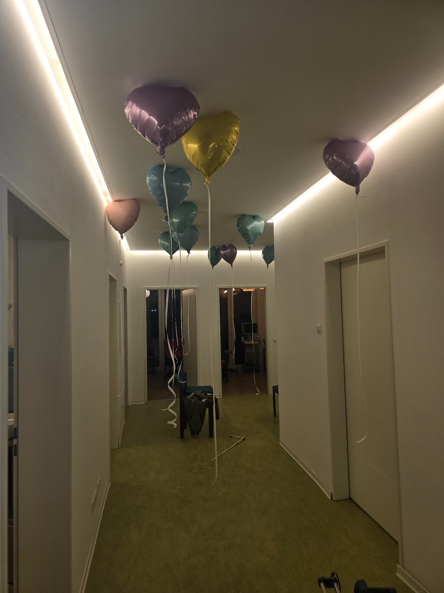 Korytarz z zieloną podłogą i białymi ścianami, oświetlony ledami, udekorowany balonami w kształcie serc w różnych kolorach, widok w perspektywie.