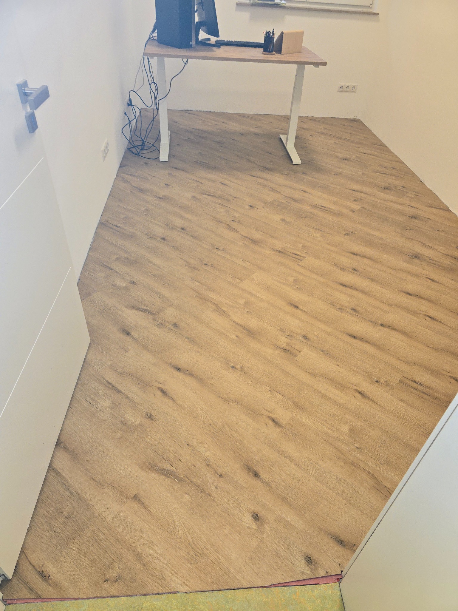 Ukończona podłoga z paneli w jasnym odcieniu drewna, ułożona diagonalnie w pokoju z białymi ścianami i biurkiem. Widoczne kable od komputera.