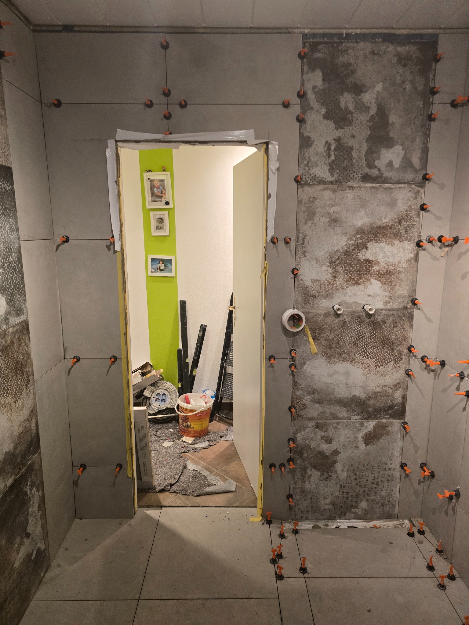 Ściana łazienki w trakcie układania szarych płytek imitujących kamień, z użyciem klipsów poziomujących. Widoczne fragmenty instalacji i narzędzia glazurnicze.