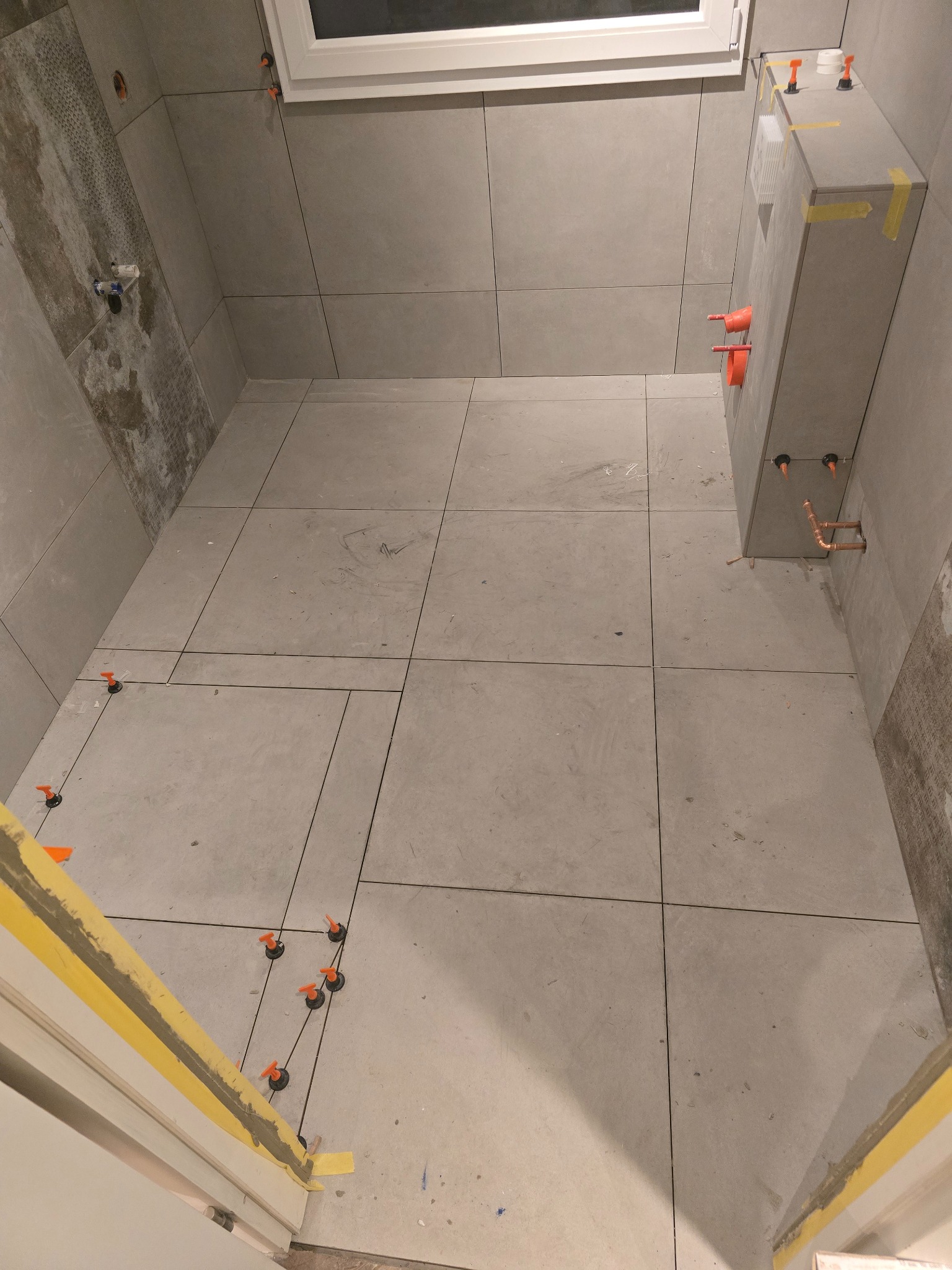 Szare płytki podłogowe i ścienne w trakcie układania w łazience, widoczne krzyżyki dystansowe i fragment instalacji podtynkowej.