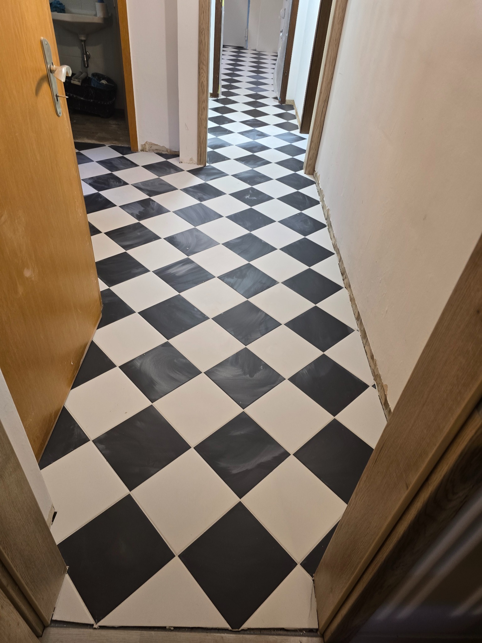 Ukończona podłoga w korytarzu z czarno-białych, kwadratowych płytek ułożonych w karo. Widoczne niedokończone ściany i futryny drzwi.