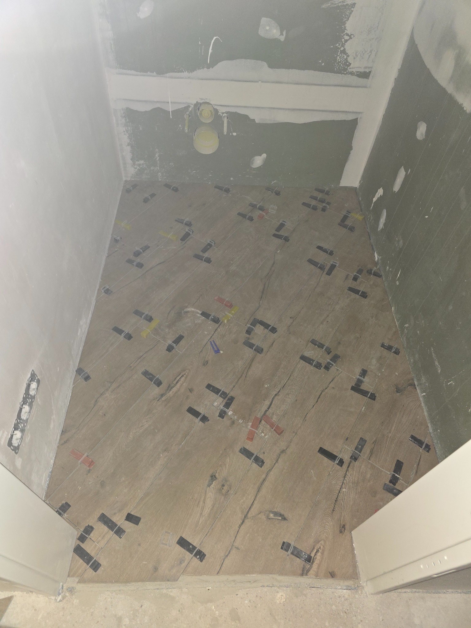 Układanie płytek imitujących drewno w małej łazience. Widoczne krzyżyki dystansowe i poziomowanie. Ściany przygotowane pod wykończenie, sufit z instalacją.