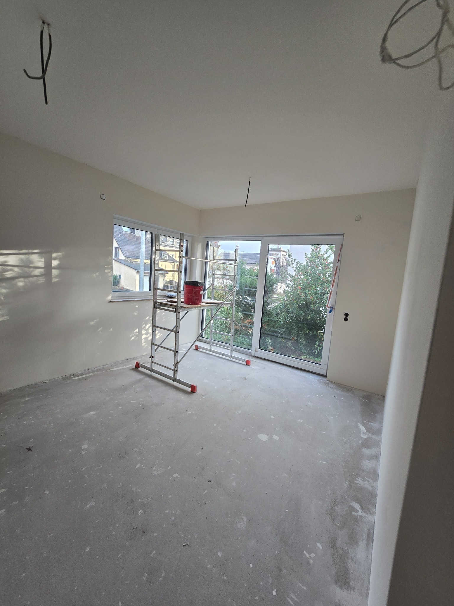 Pokój w trakcie prac wykończeniowych: ściany po gładziowaniu, widoczne rusztowanie z wiadrem farby, surowa podłoga. Przygotowanie do malowania.