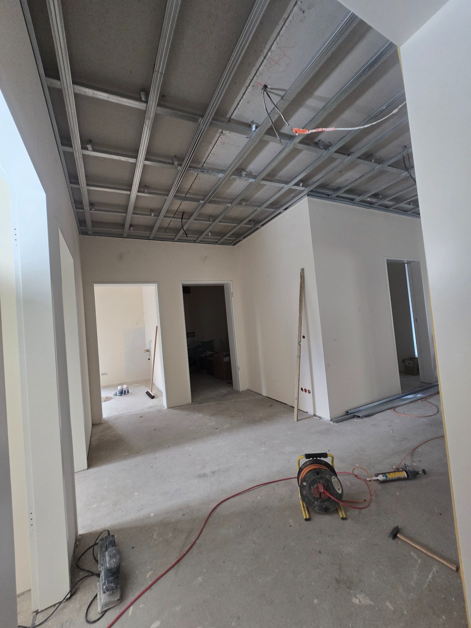 Montaż sufitu podwieszanego z płyt karton-gips na metalowej konstrukcji w jasnym wnętrzu. Widoczne narzędzia i kable. W tle otwarte przejścia do innych pomieszczeń.