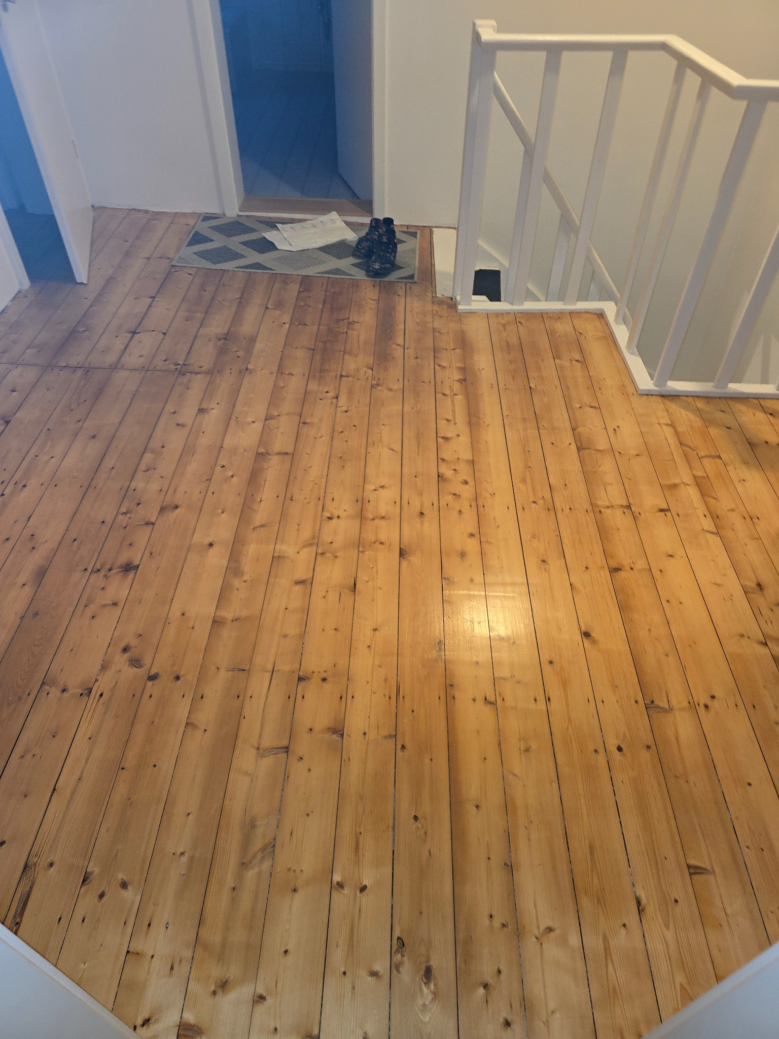 Wykończona podłoga z desek w odcieniach brązu, widok z góry, z fragmentem białych schodów i dywanikiem przy drzwiach. Naturalne drewno z widocznymi słojami.