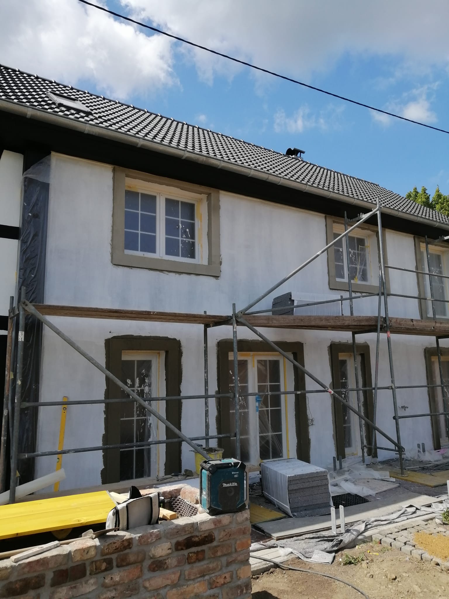 Elewacja domu w trakcie prac: nałożony tynk wokół okien, rusztowania, materiały budowlane. Widoczne przygotowania do dalszych etapów prac wykończeniowych.