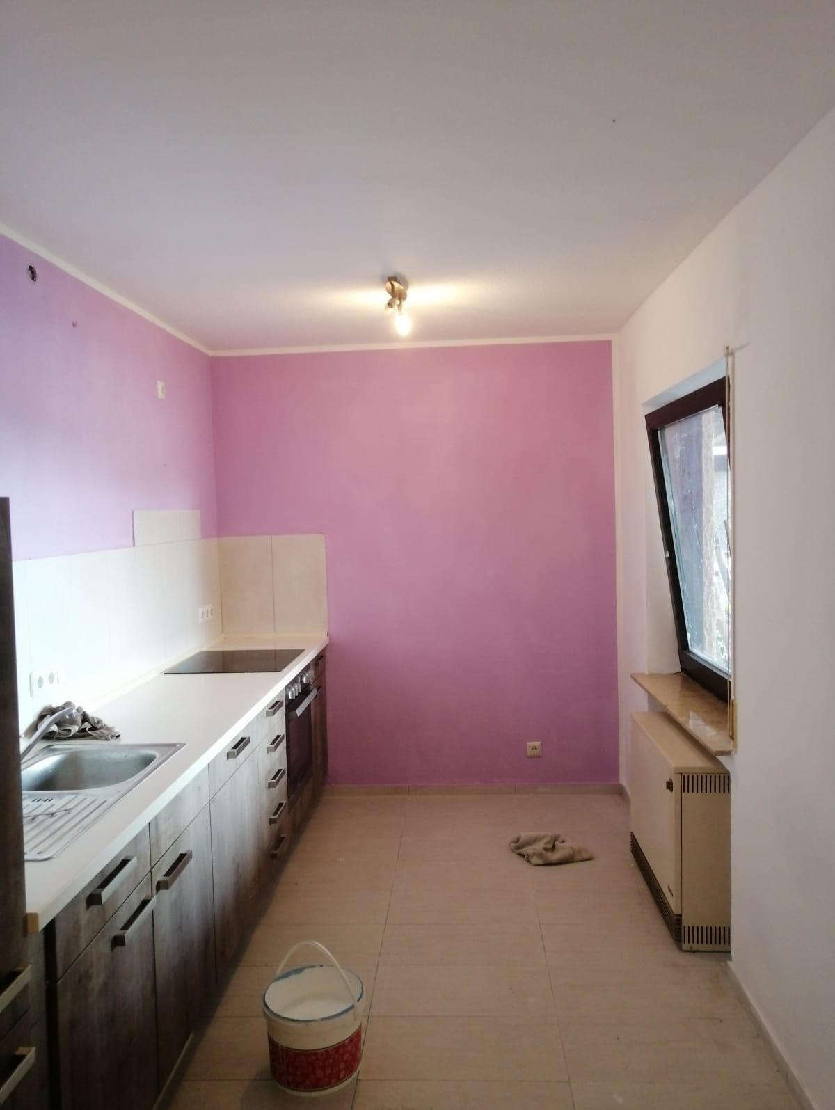 Kuchnia po malowaniu ścian na fioletowo, widoczny fragment zabudowy kuchennej, okno i grzejnik. Na podłodze wiadro z farbą i szmata. Ściana pomalowana na gładko.
