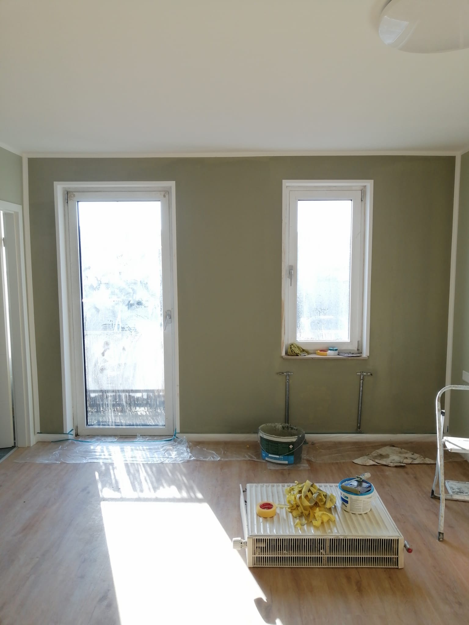Pokój po malowaniu ścian na oliwkowy kolor, z widocznymi narzędziami malarskimi na grzejniku i zabezpieczeniami pod oknami. Świeżo pomalowane ściany i jasna podłoga.