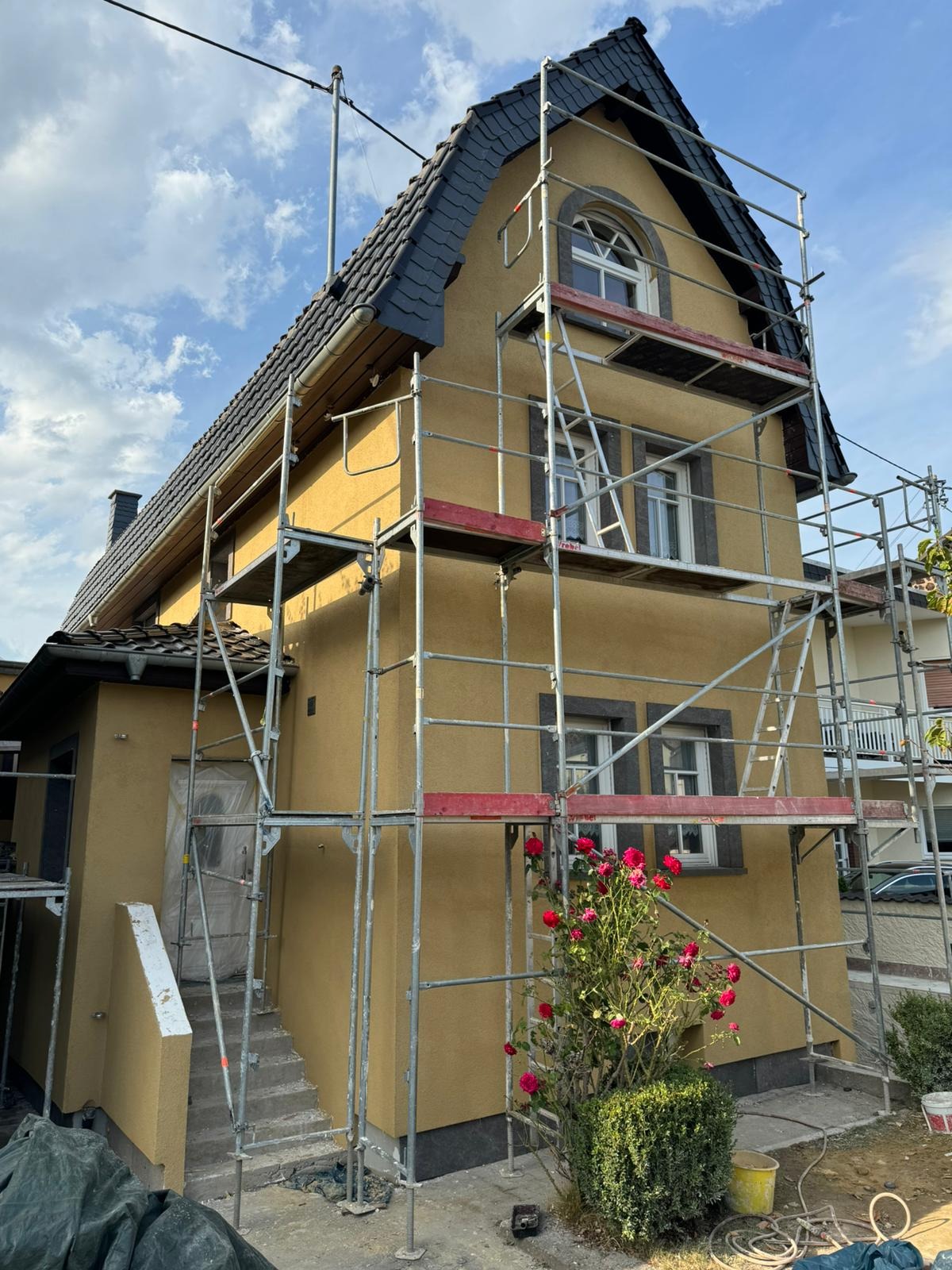 Dom w trakcie ocieplania elewacji, otoczony rusztowaniami. Widoczne róże w ogrodzie i fragment nieba. Elewacja w odcieniach żółci, dach kryty ciemną dachówką.