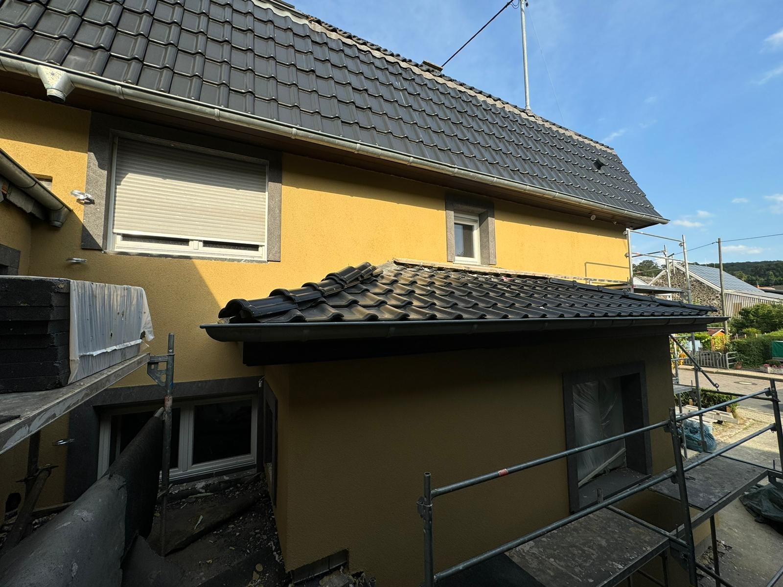 Żółty dom z czarnym dachem w trakcie prac elewacyjnych, widoczne rusztowania i okno z roletą. Ujęcie z dołu, fragment dachu drugiego budynku w tle.