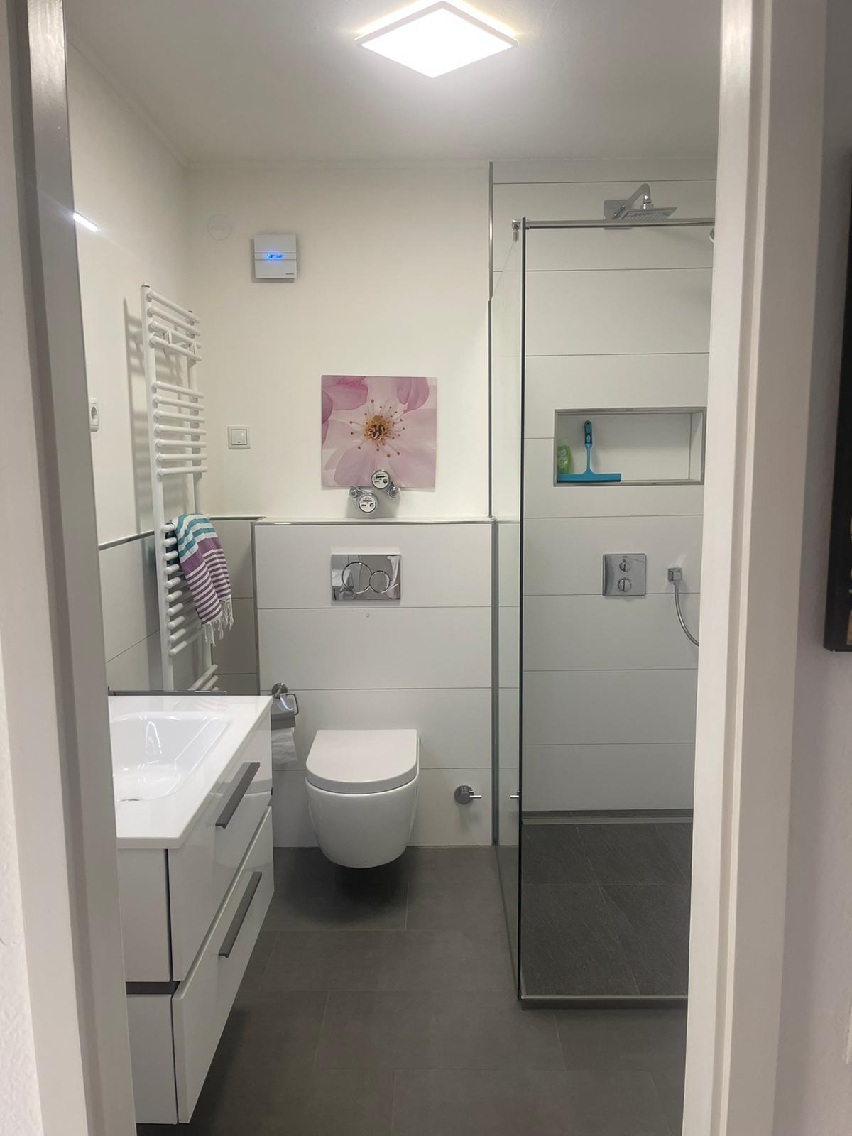 Nowoczesna łazienka z kabiną prysznicową typu walk-in, umywalką z szafką, podwieszaną toaletą i grzejnikiem drabinkowym. Minimalistyczny design, szare płytki podłogowe.