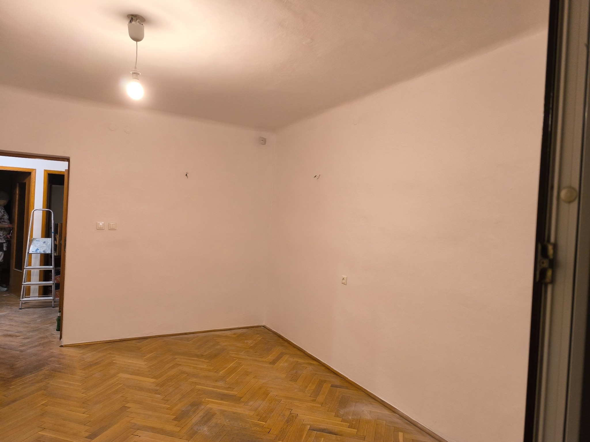 Jasne wnętrze po remoncie: świeżo pomalowane ściany w odcieniu beżu, parkiet w jodełkę, drabina w tle. Widoczne gniazdka i punkty po demontażu elementów dekoracyjnych.