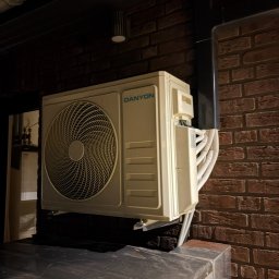 DKV INSTAL - Zamontowany klimatyzator Danyon na ceglanej ścianie w Otyniu. Widoczne rury odprowadzające. Oświetlenie punktowe podkreśla fakturę ściany i urządzenie.