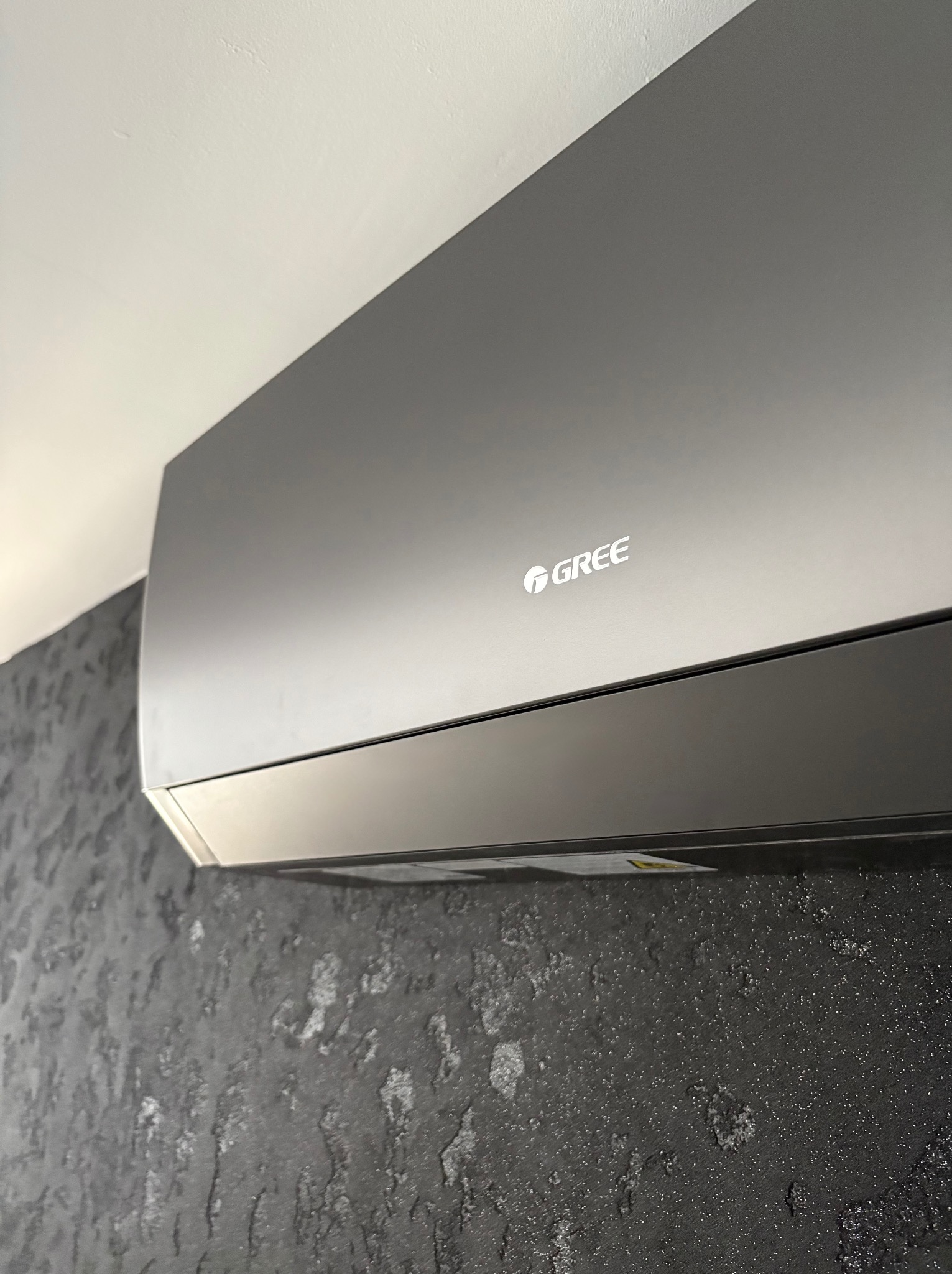Szary klimatyzator marki Gree zamontowany na ciemnej, teksturowanej ścianie. Widoczny fragment sufitu. Ujęcie z dołu, podkreślające nowoczesny design urządzenia.
