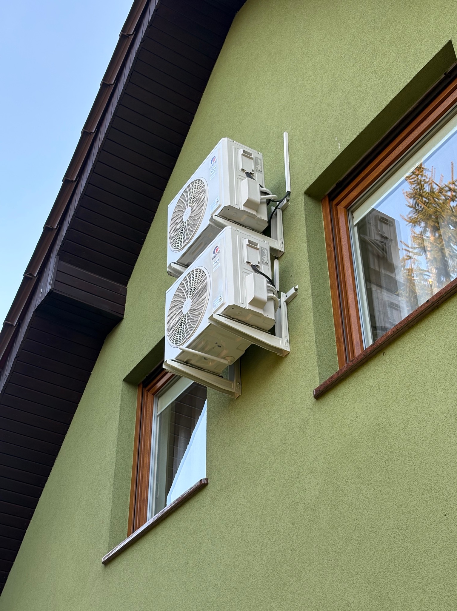 Dwa białe klimatyzatory zamontowane jeden nad drugim na zielonej ścianie domu, widoczne okna z drewnianymi ramami i fragment dachu.