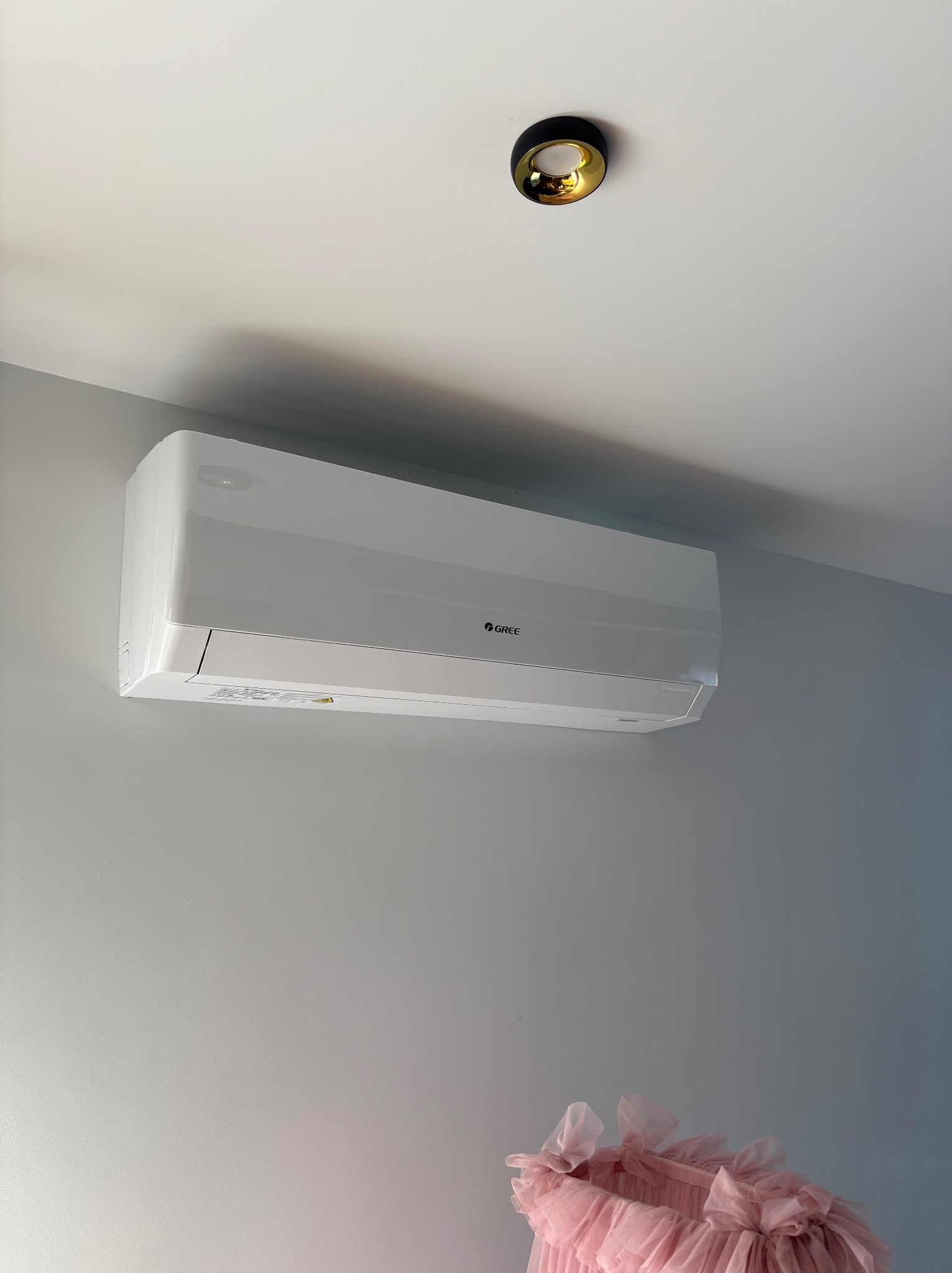 Biały klimatyzator Gree na szarej ścianie, pod złotą lampą sufitową. Fragment różowej dekoracji w dolnym rogu kadru. Ujęcie z dołu, podkreślające minimalistyczny design.