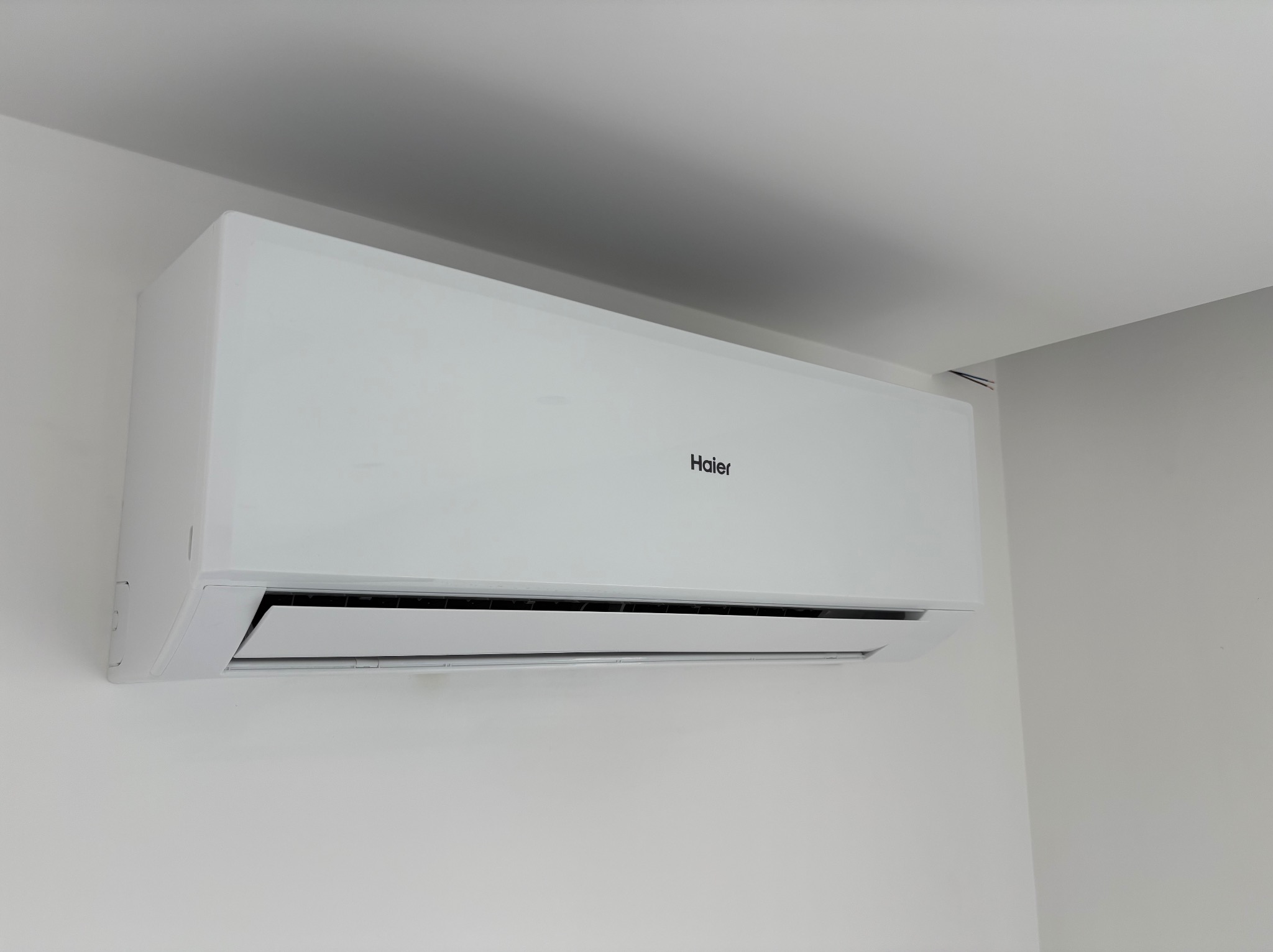 Biała jednostka klimatyzacji Haier zamontowana na jasnej ścianie, widoczna z boku, z delikatnym cieniem na suficie w rogu pomieszczenia.
