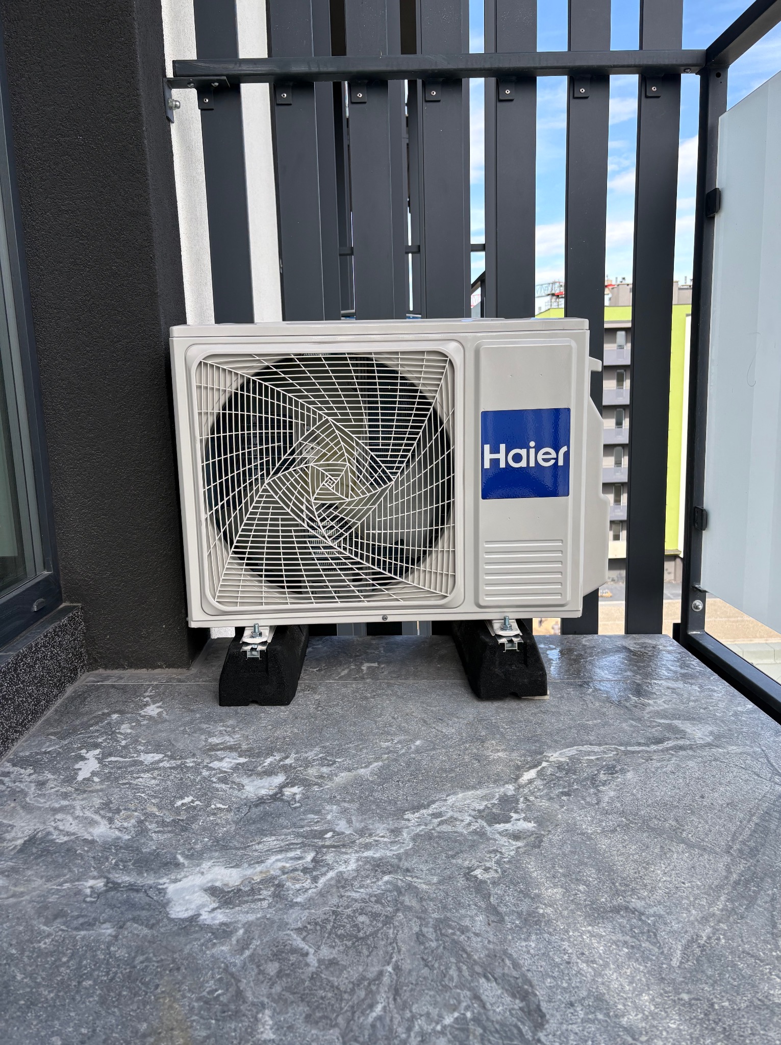 Biała jednostka zewnętrzna klimatyzacji Haier na balkonie, zamontowana na czarnych podkładkach antywibracyjnych, z widokiem na błękitne niebo przez metalową balustradę.
