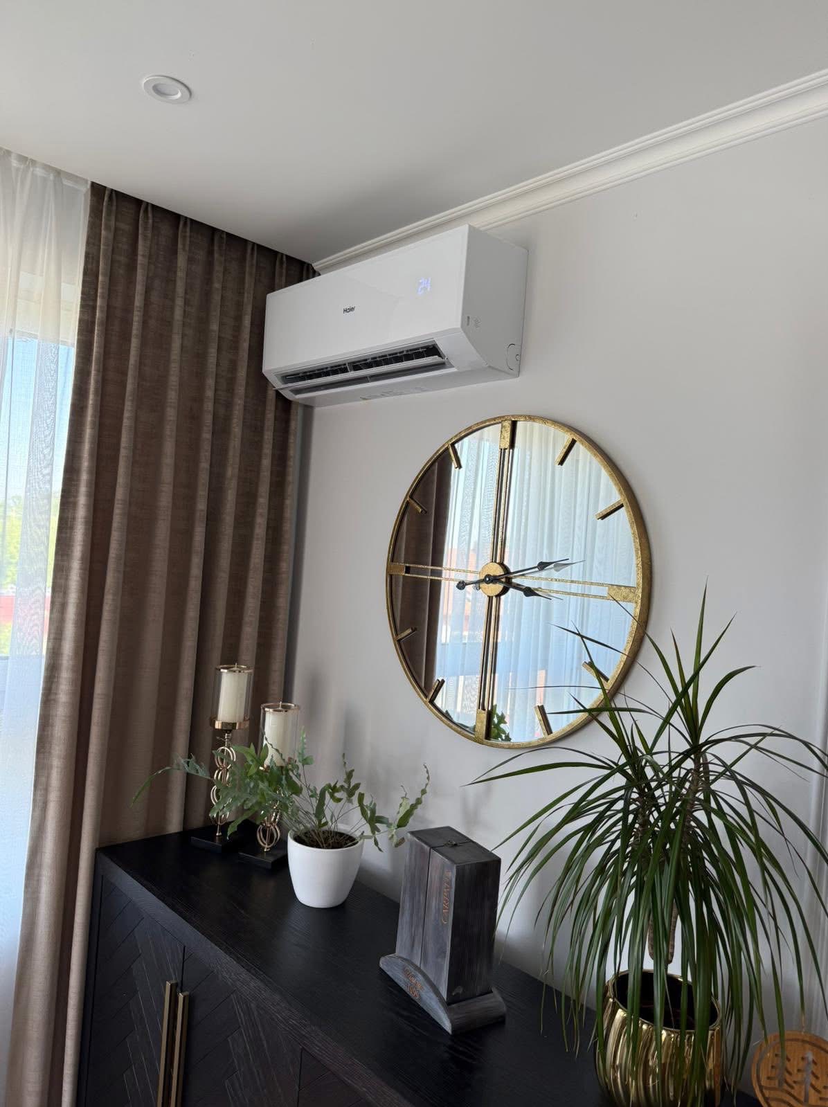 Biała klimatyzacja Haier na ścianie nad komodą z dekoracjami i zegarem w złotym obramowaniu. Widok na okno z brązowymi zasłonami.