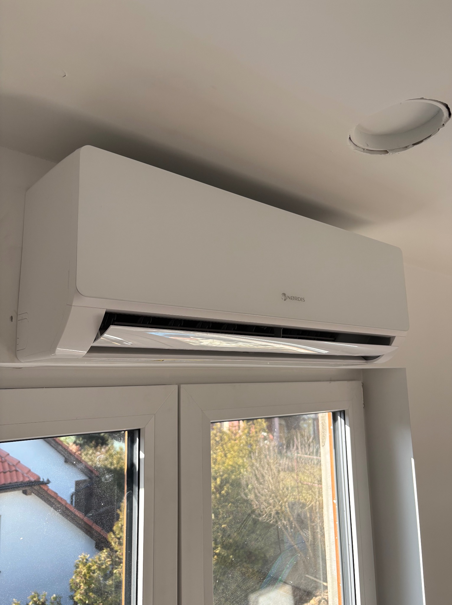 Biały klimatyzator NORDIS zamontowany nad oknem, widok z dołu. Na suficie widoczny otwór wentylacyjny. Widok na zewnątrz przez okno.