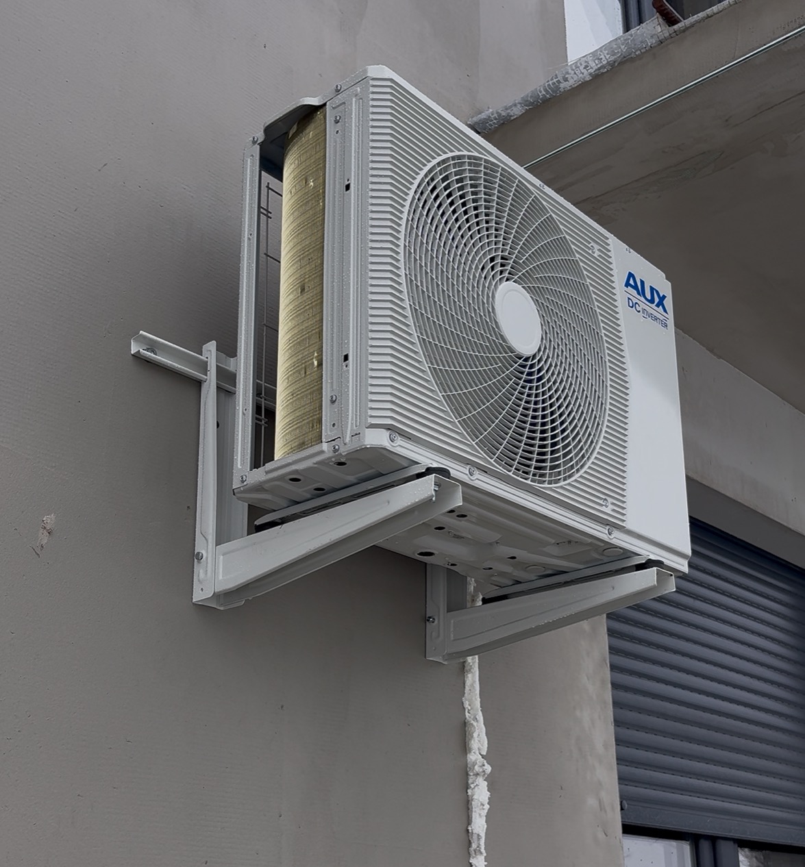 Biała jednostka zewnętrzna klimatyzacji AUX DC Inverter zamontowana na szarej ścianie budynku, widoczne elementy montażowe i izolacja. Ujęcie z dołu.