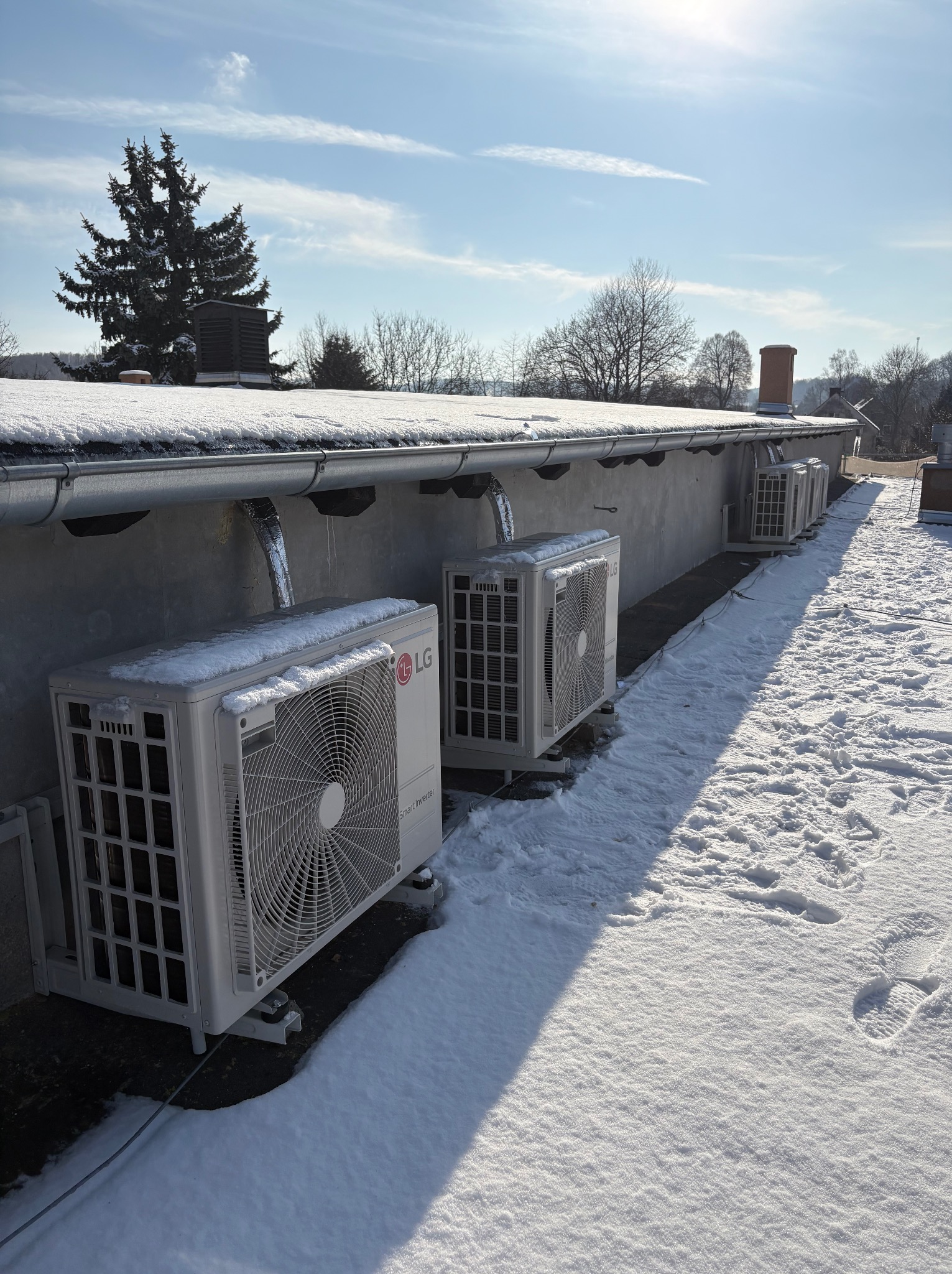 Rząd białych jednostek zewnętrznych klimatyzacji LG na dachu pokrytym śniegiem, z widocznymi przewodami i elementami montażowymi. Błękitne niebo w tle.