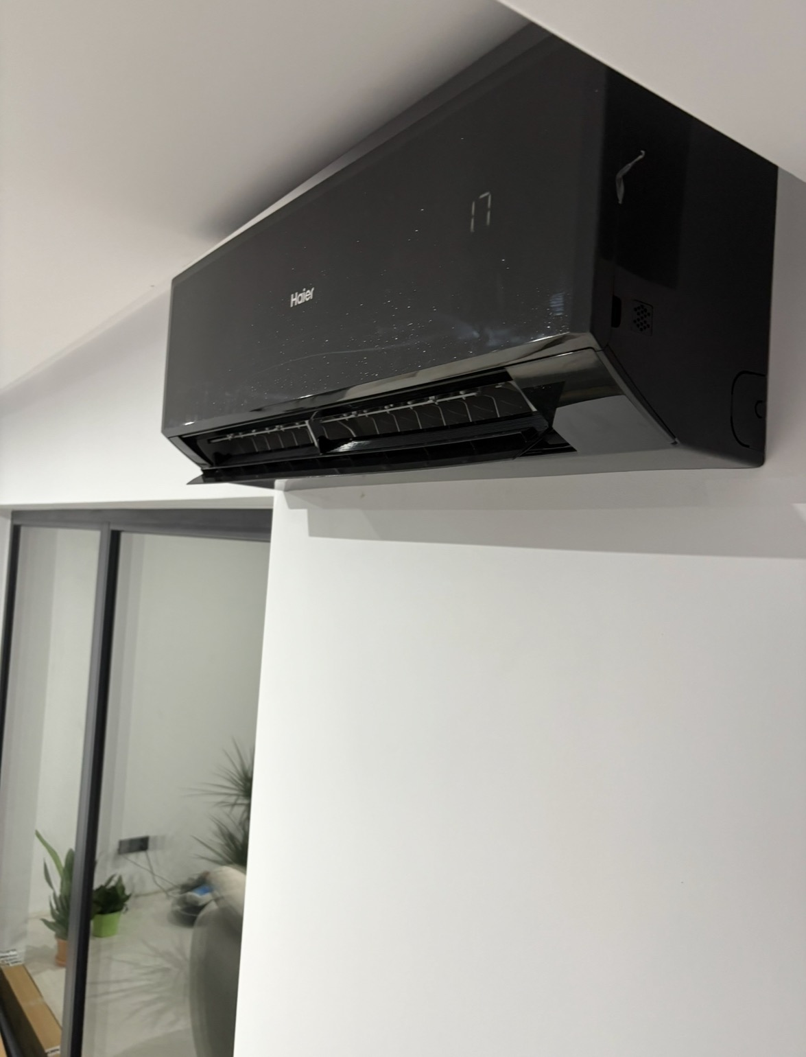 Czarny klimatyzator Haier zamontowany na białej ścianie, widoczny z boku, z ustawioną temperaturą 17 stopni. W tle okno z widokiem na wnętrze.