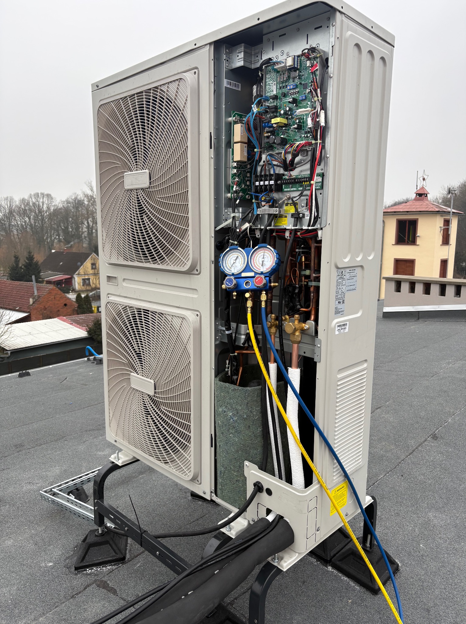 Otwarta obudowa klimatyzatora na dachu, widoczne podzespoły, manometry i przewody serwisowe. W tle budynki mieszkalne i pochmurne niebo.