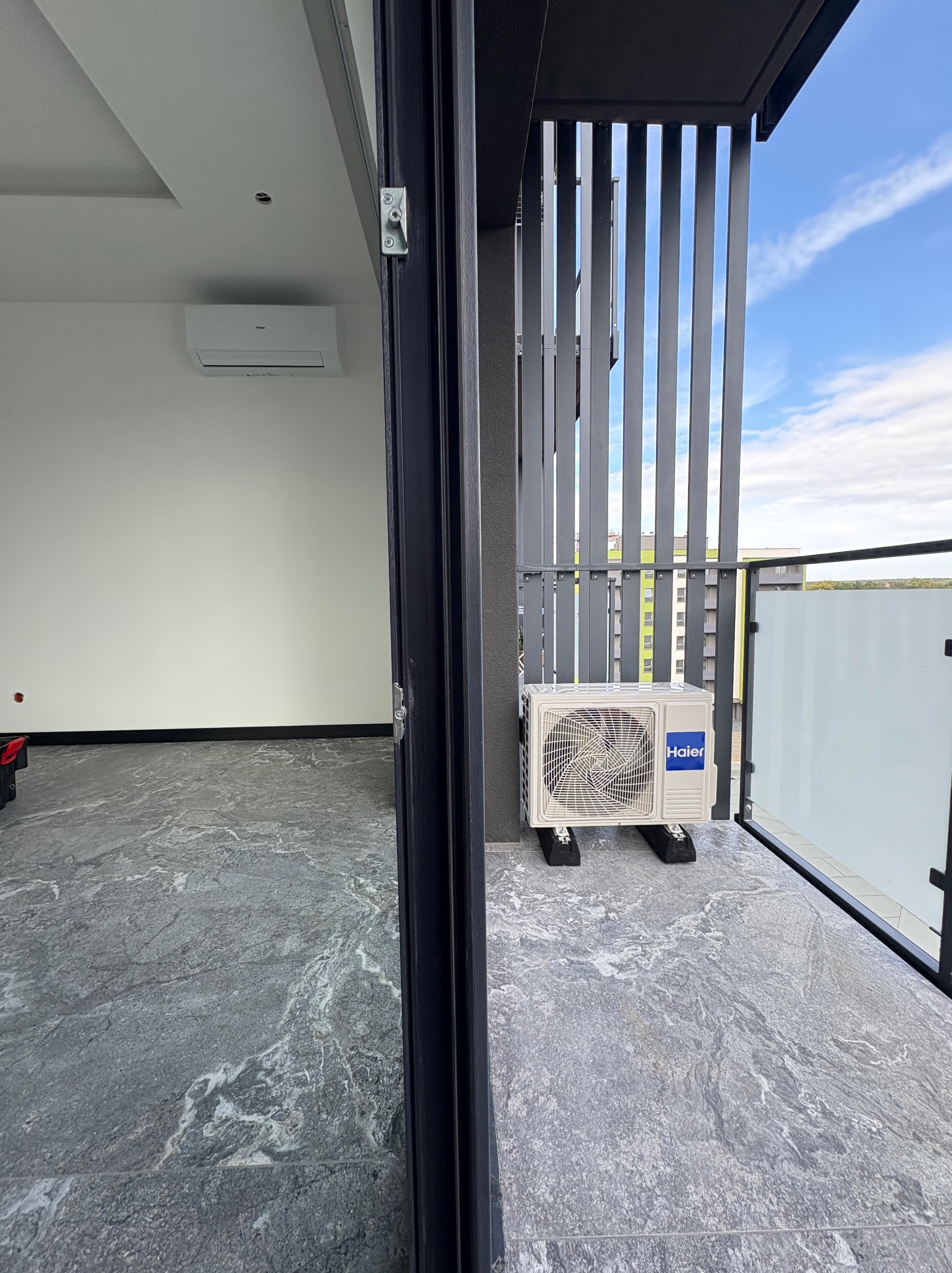 Zamontowany klimatyzator Haier na balkonie w Otyniu. Widoczna jednostka zewnętrzna na tle błękitnego nieba i fragment wnętrza z jednostką wewnętrzną. Szare płytki podłogowe.