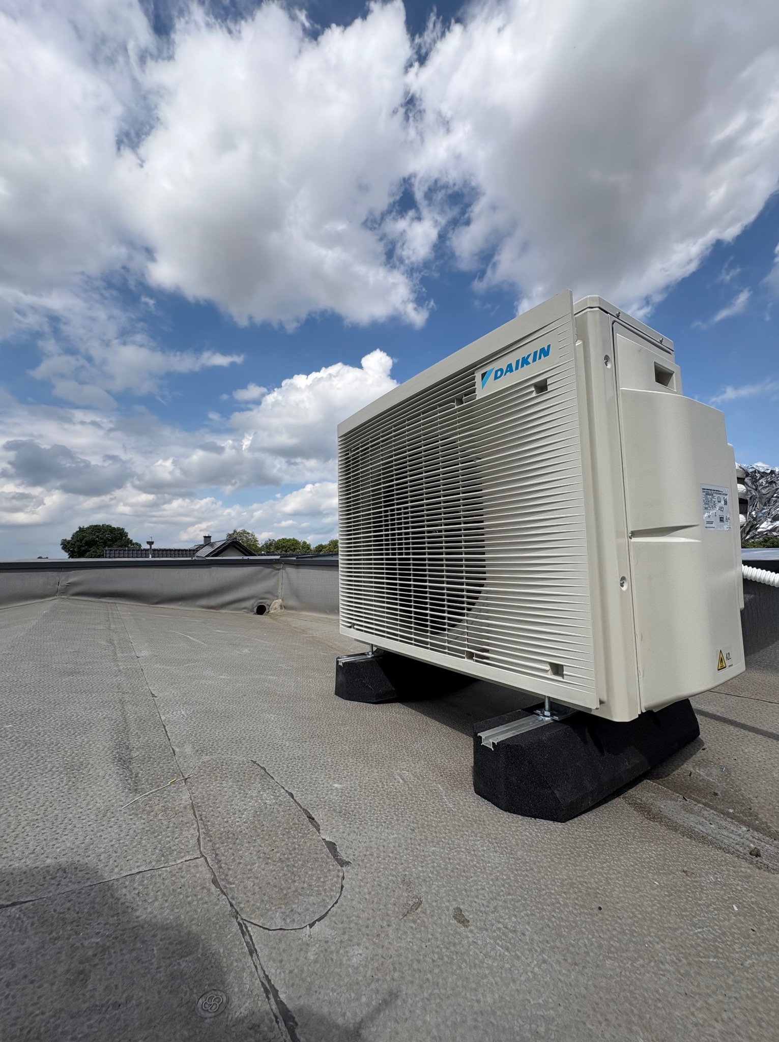 Biała jednostka zewnętrzna klimatyzacji Daikin na dachu, zamontowana na czarnych podkładkach antywibracyjnych. W tle błękitne niebo z obłokami.