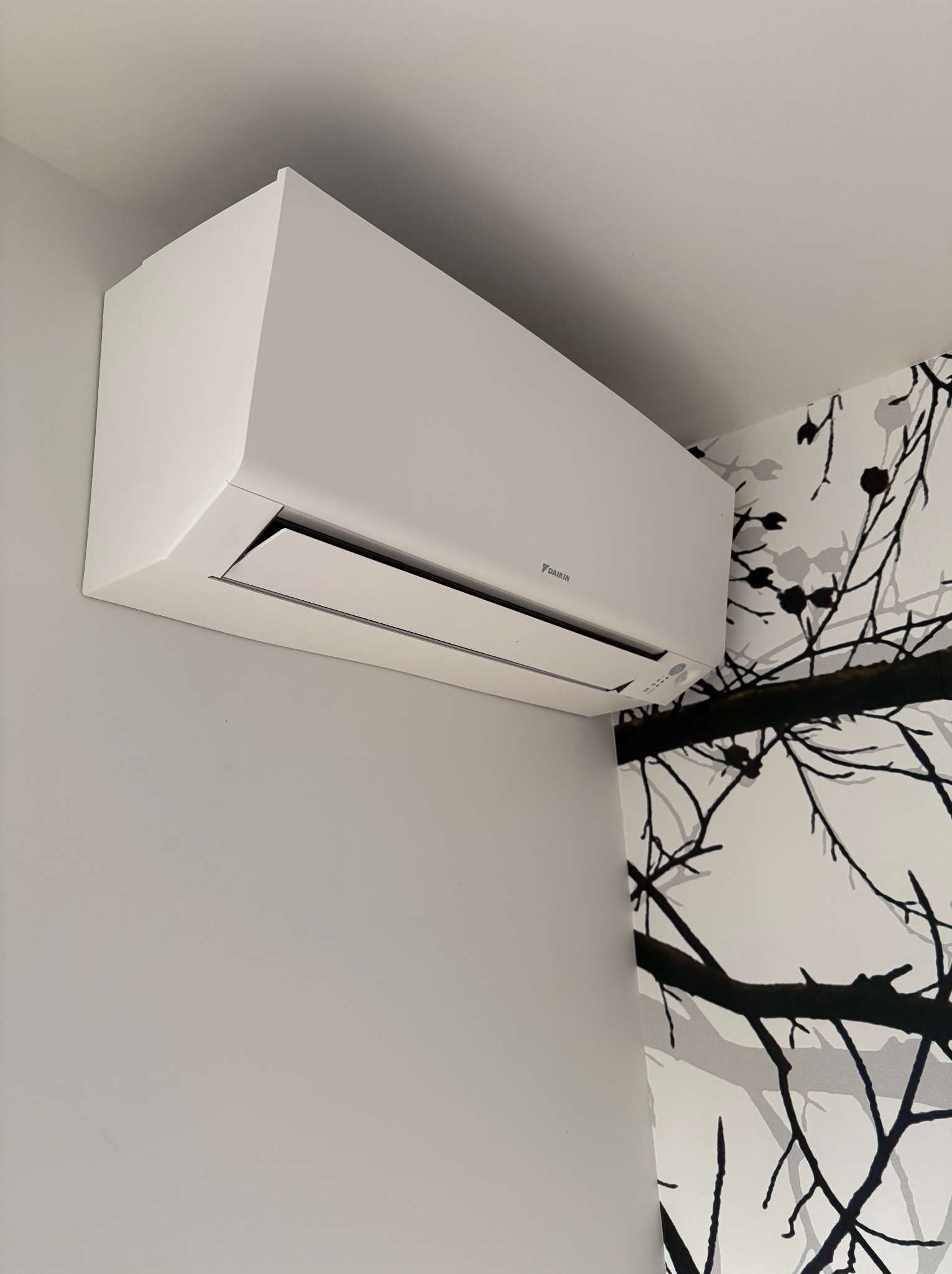 Biała jednostka klimatyzacji Daikin zamontowana na ścianie z szarą farbą, obok ściany z tapetą w czarno-białe gałęzie. Minimalistyczny design.