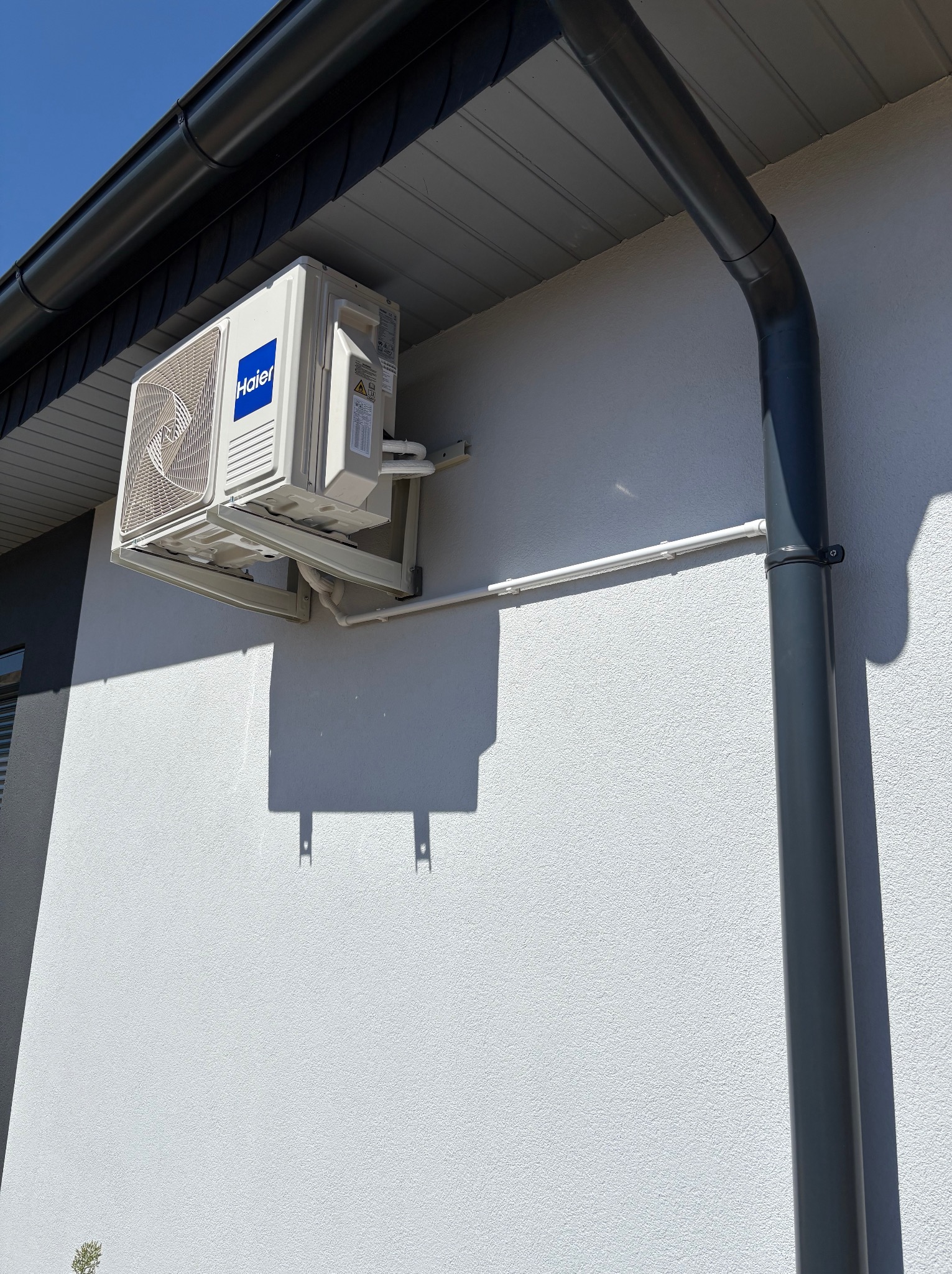 Biała jednostka zewnętrzna klimatyzacji Haier zamontowana na elewacji budynku z szarym orynnowaniem i białą rurką odprowadzającą skropliny.