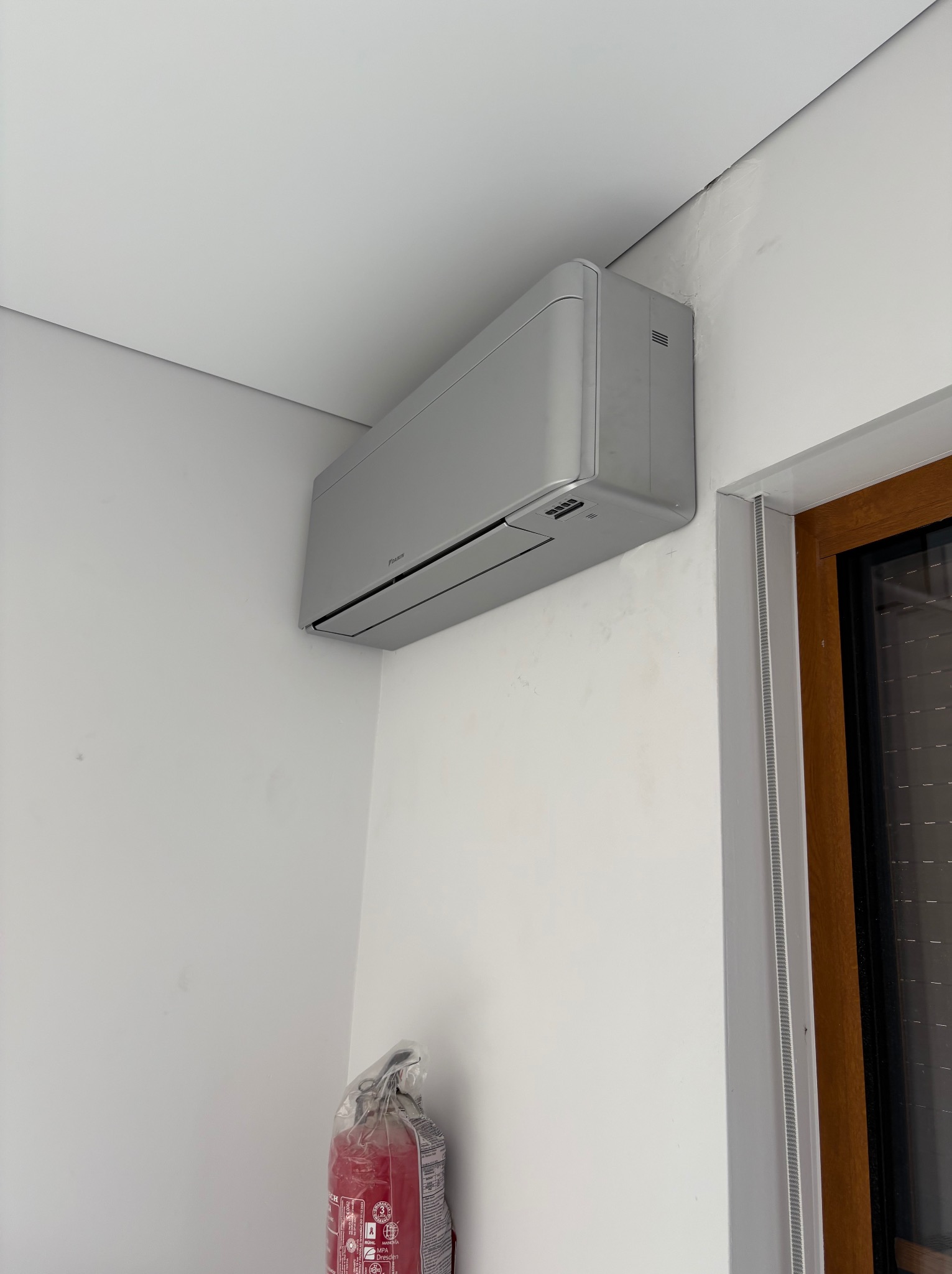 Szary klimatyzator Daikin zamontowany na białej ścianie, widoczny fragment okna i gaśnica w folii. Minimalistyczny design, jasne wnętrze.