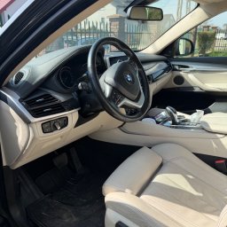 Auto Spa - Jasne skórzane wnętrze BMW X6 z widocznymi detalami deski rozdzielczej, kierownicy i foteli. Widok z zewnątrz auta, w tle ogrodzenie i dom.